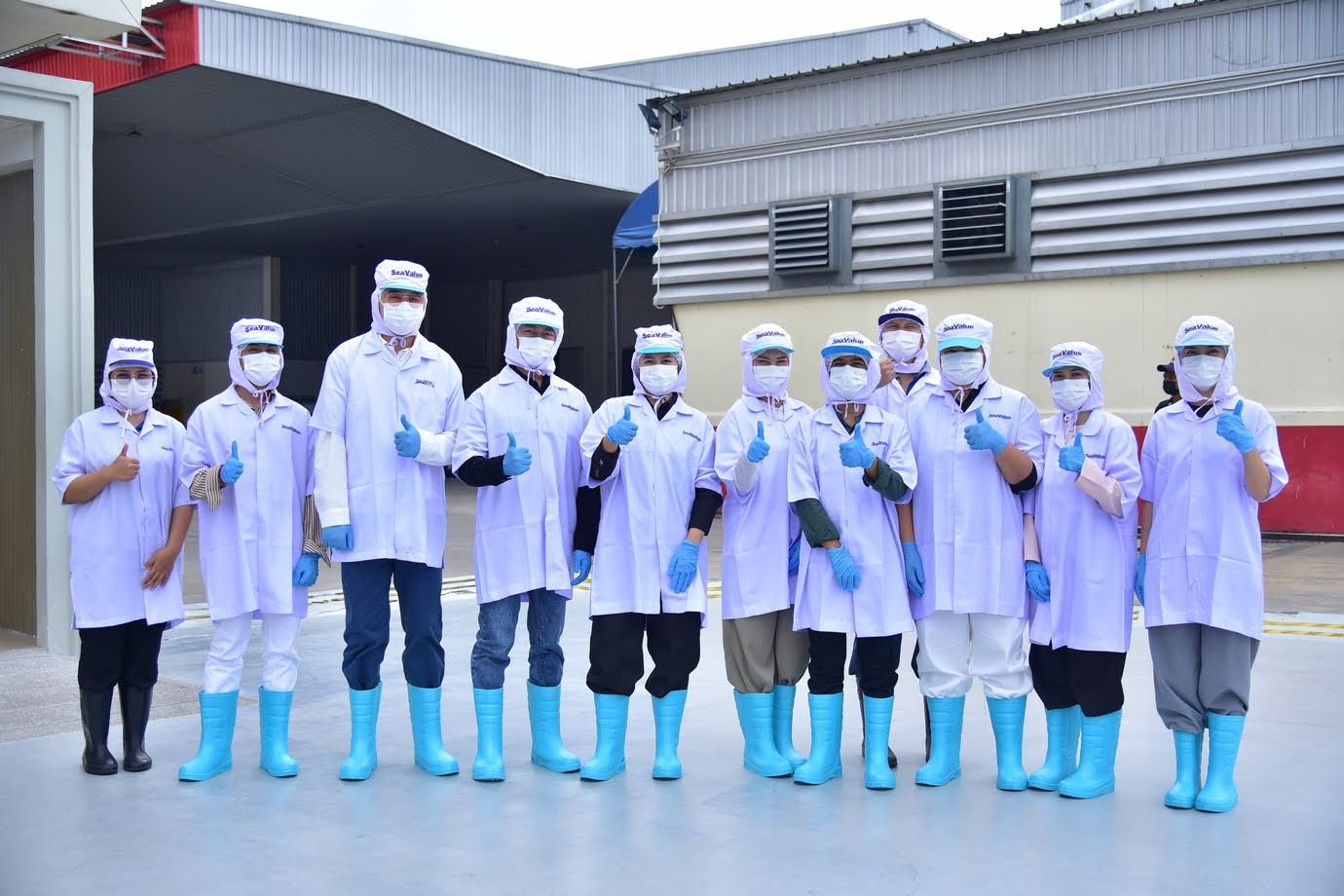Workers in white coats, masks, and blue boots give thumbs-up outside a factory.