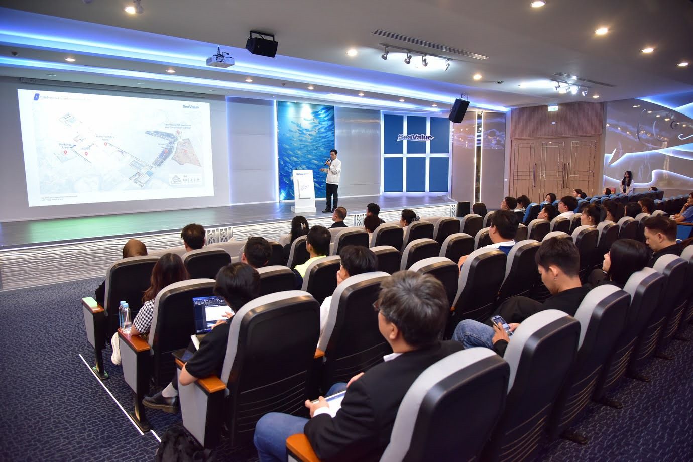 Speaker presents to an audience in a modern conference room with a large screen.