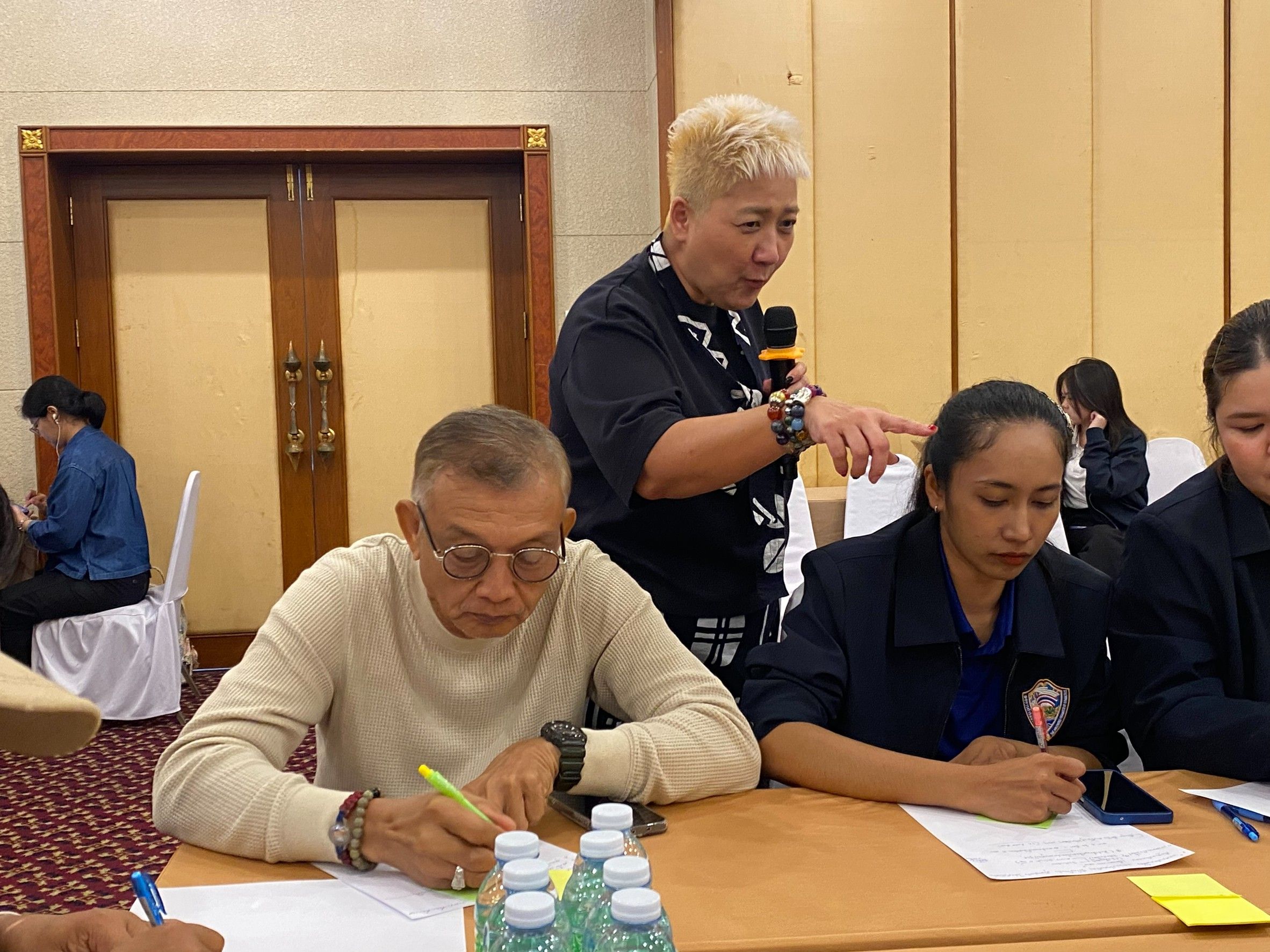 Woman speaking into microphone while people write at a workshop table.