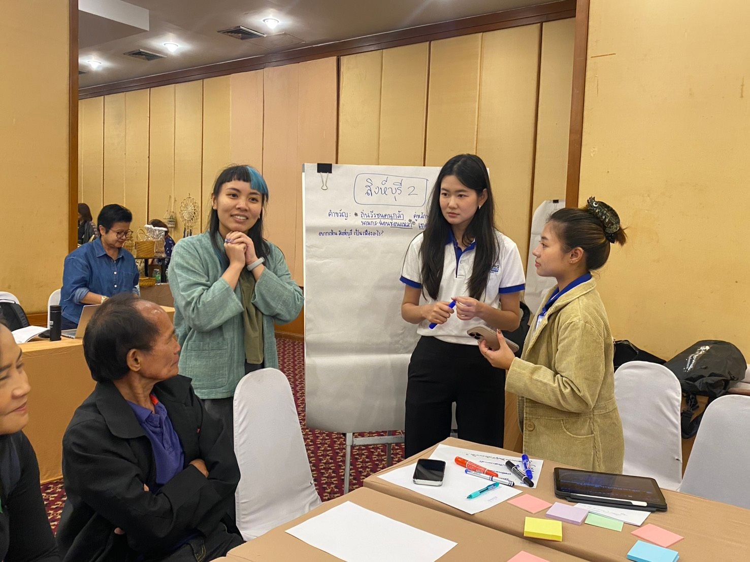 Three women present at a whiteboard during a meeting, while another listens attentively.