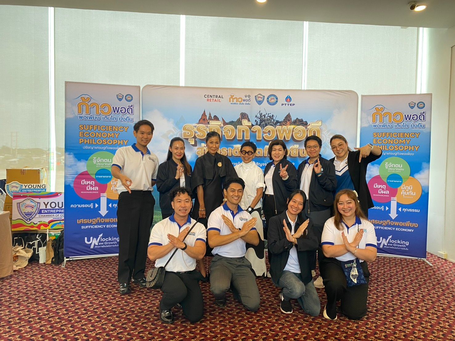 Group of smiling people making a traditional Thai wai gesture in front of an event banner.