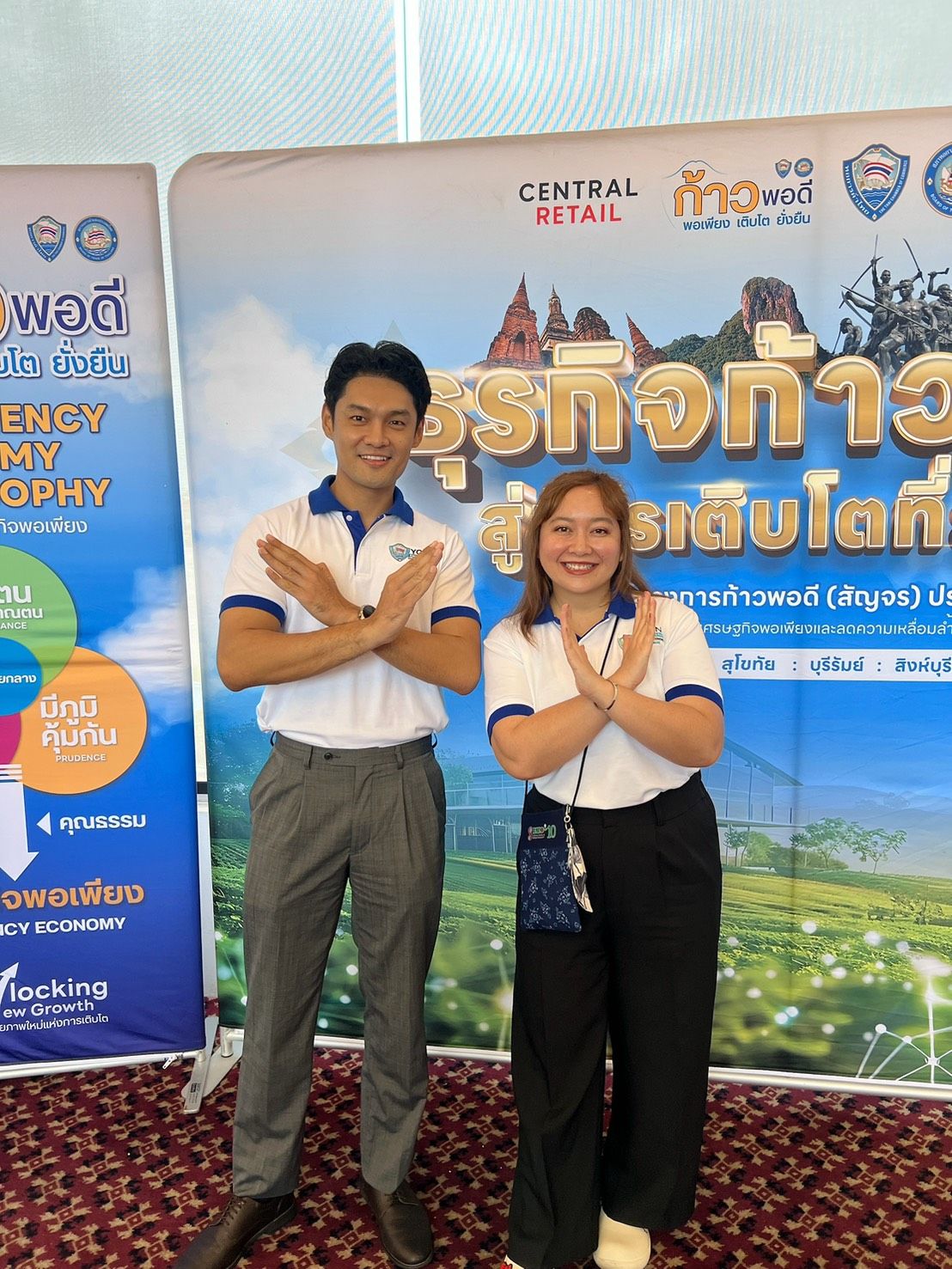 Man and woman in white shirts make crossed-arm gesture before promotional banners.