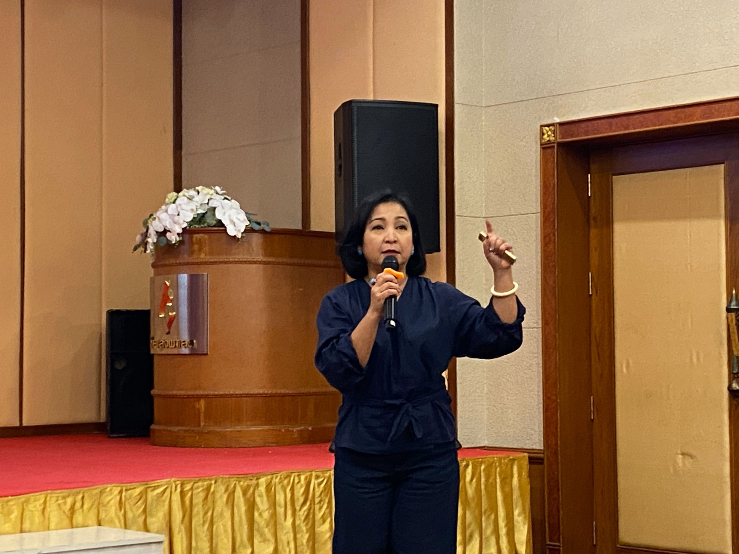 Woman on stage speaks into microphone, gesturing with raised hand during a presentation.