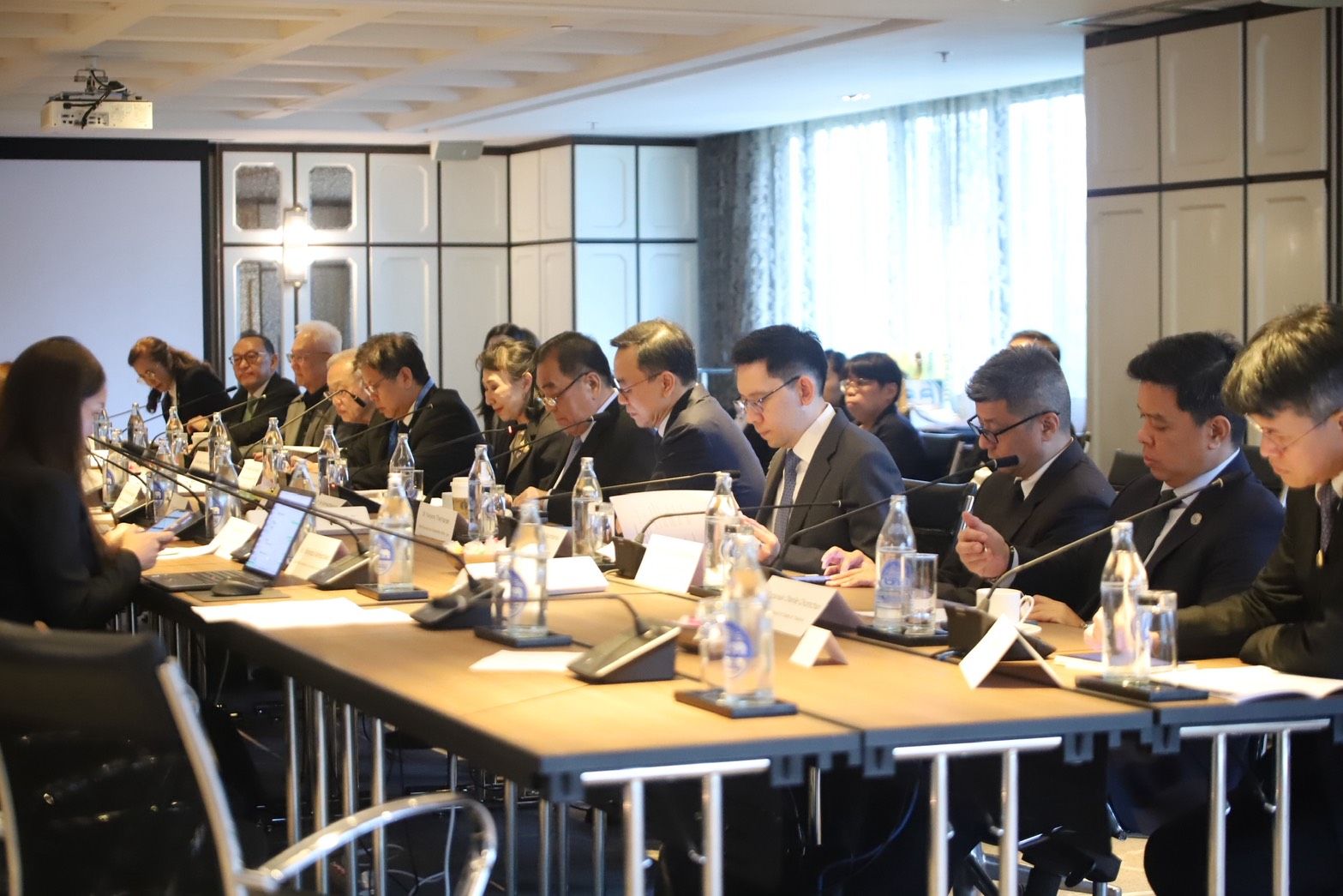 Professionals attend a formal meeting, seated around a long conference table with laptops and microphones.