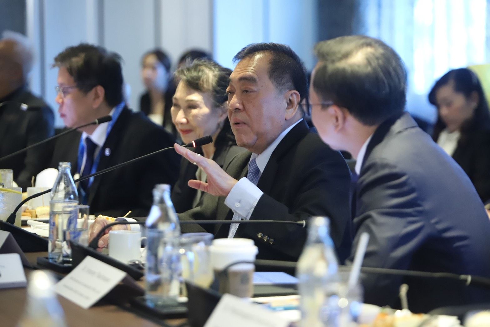 An Asian man in a suit gestures while speaking at a formal meeting table.