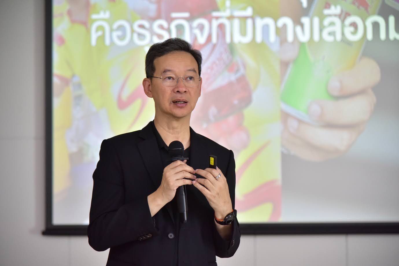 A bespectacled man in a black shirt speaks into a microphone, gesturing before a Thai presentation.