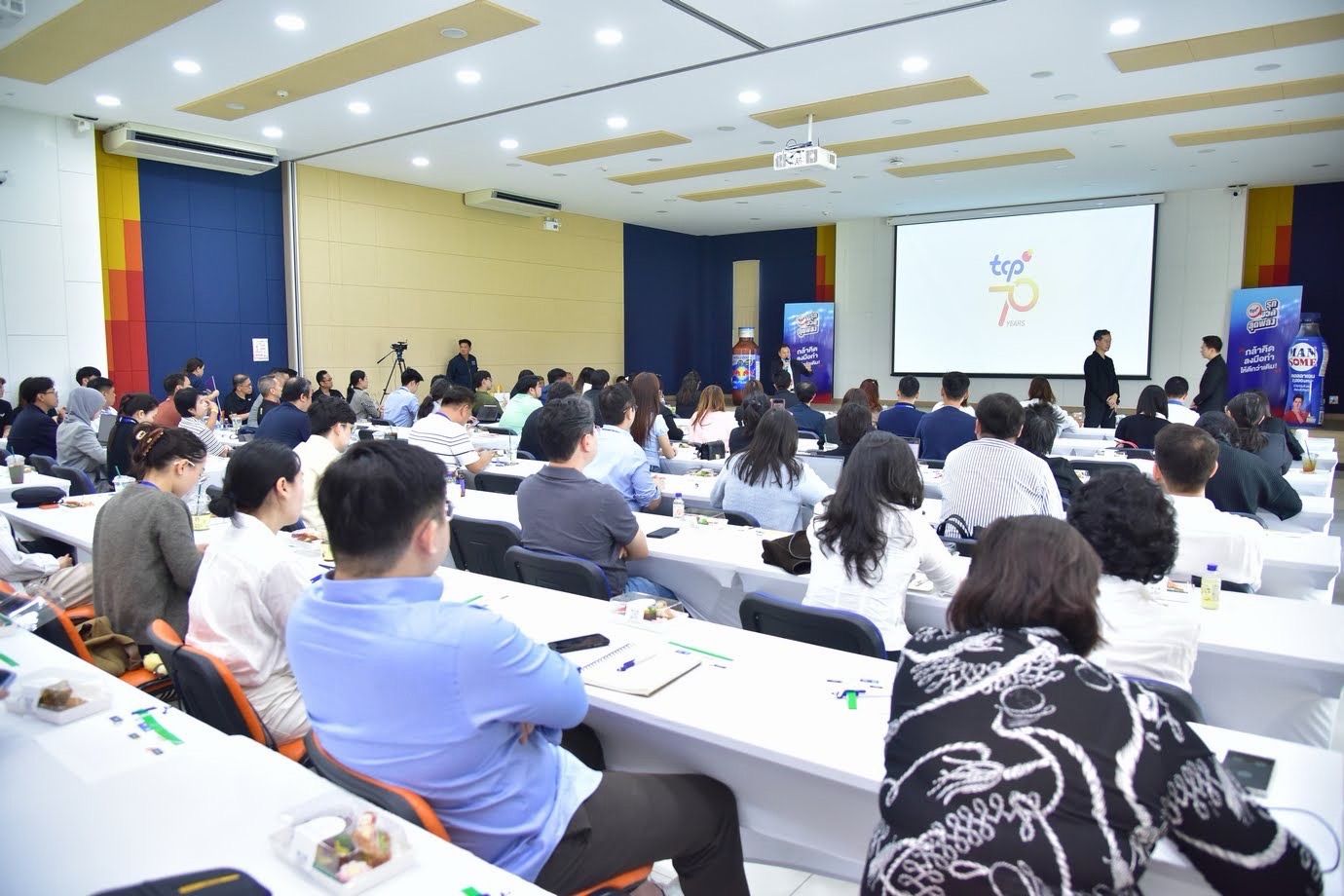 Large audience attends a conference, watching a presentation on a projector screen.