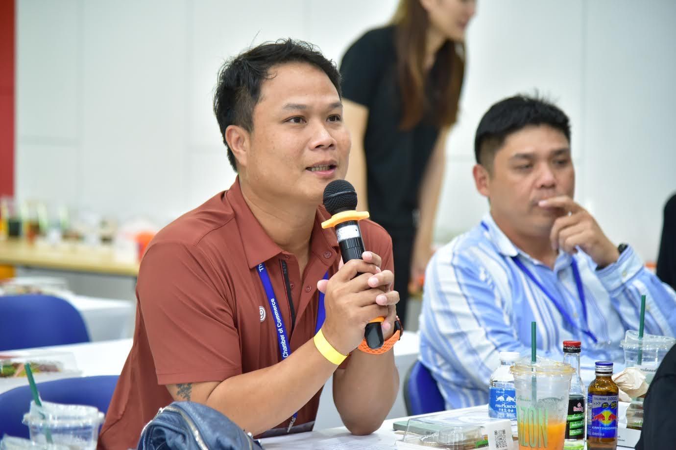 Man in a reddish-brown shirt speaks into a microphone at an indoor event.