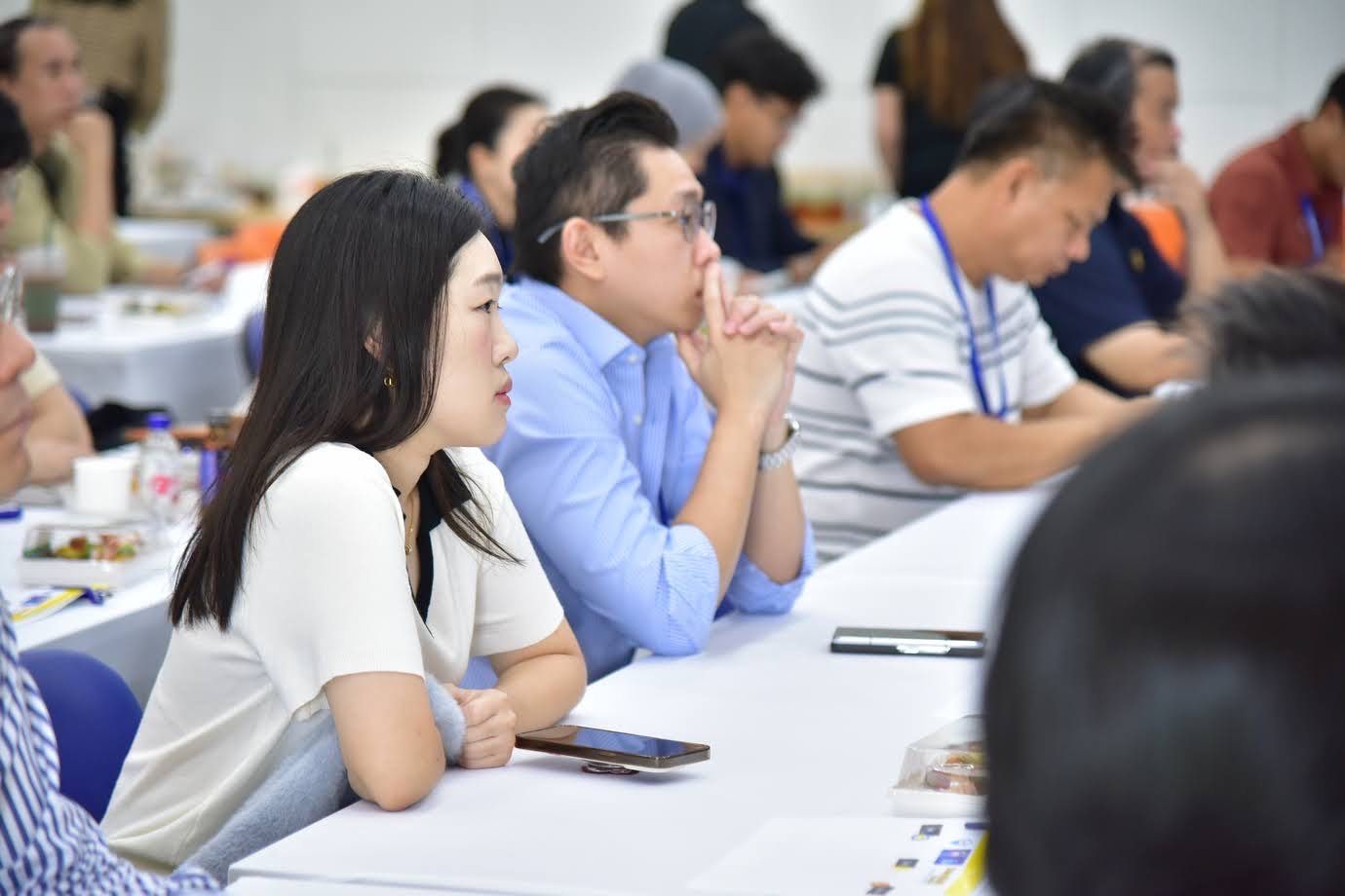 Attendees, mostly East Asian, engaged in a conference or seminar setting, listening intently.