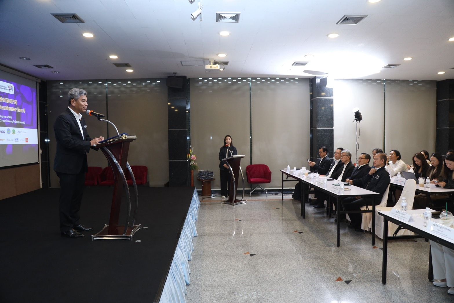 Man in a suit speaks at a podium to an audience in a conference room.