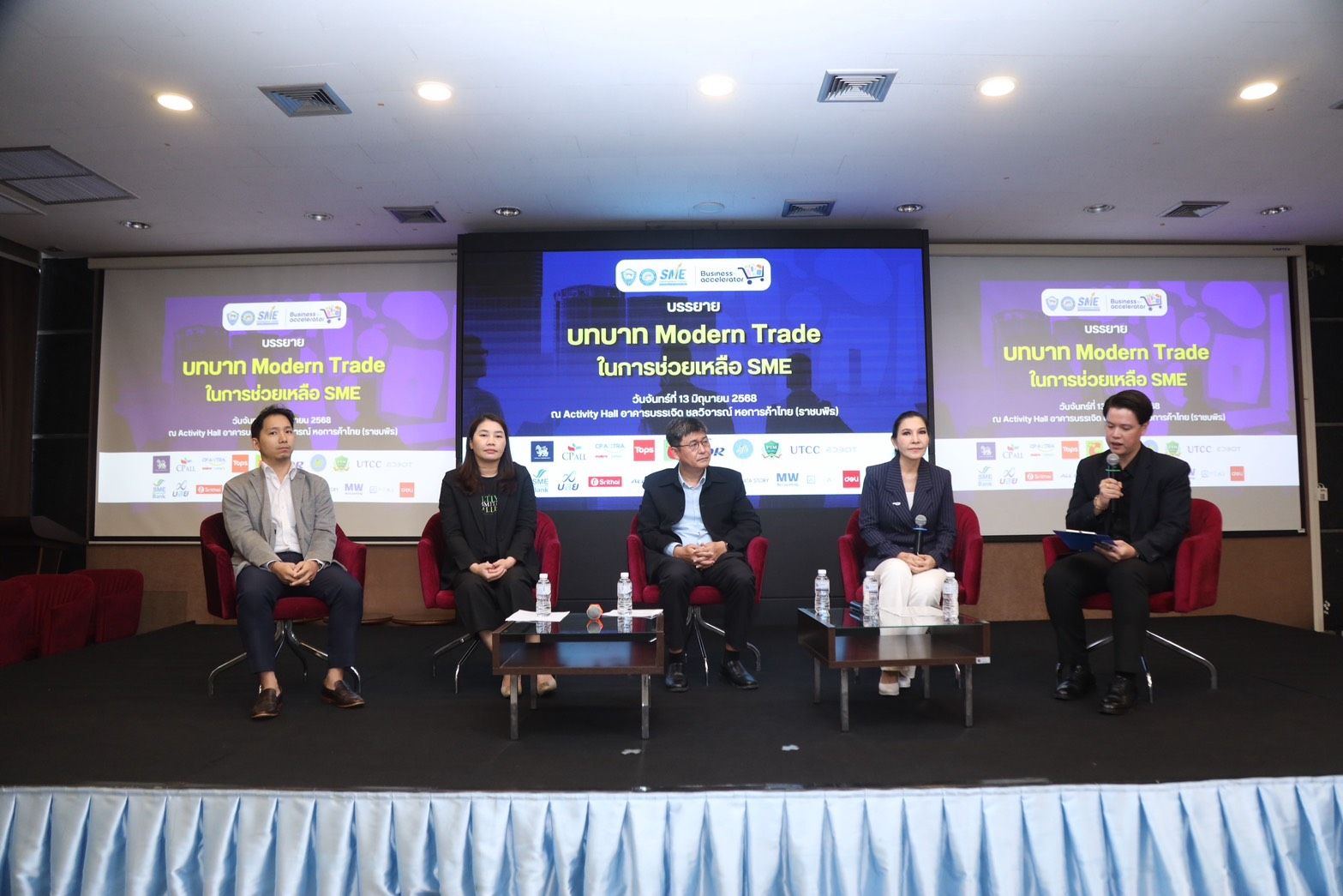 Five panelists seated on a stage during a Modern Trade conference with screens displaying event information.