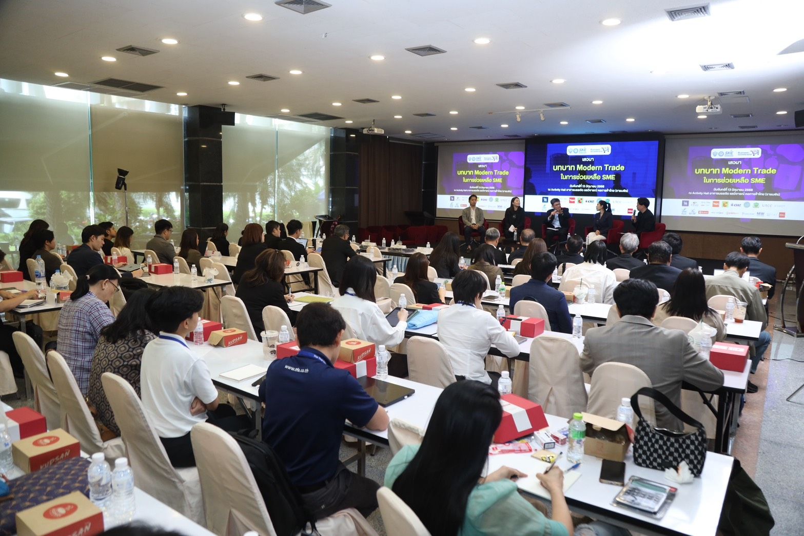 Audience seated at tables in a conference room with two large presentation screens.