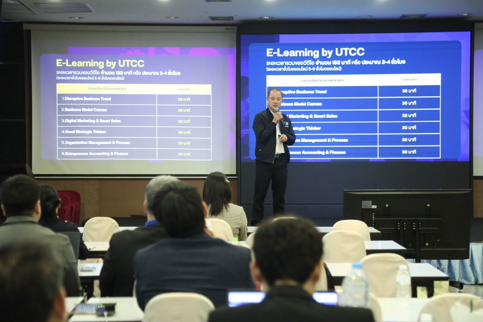 A man presents "E-Learning by UTCC" on large screens to an attentive audience.