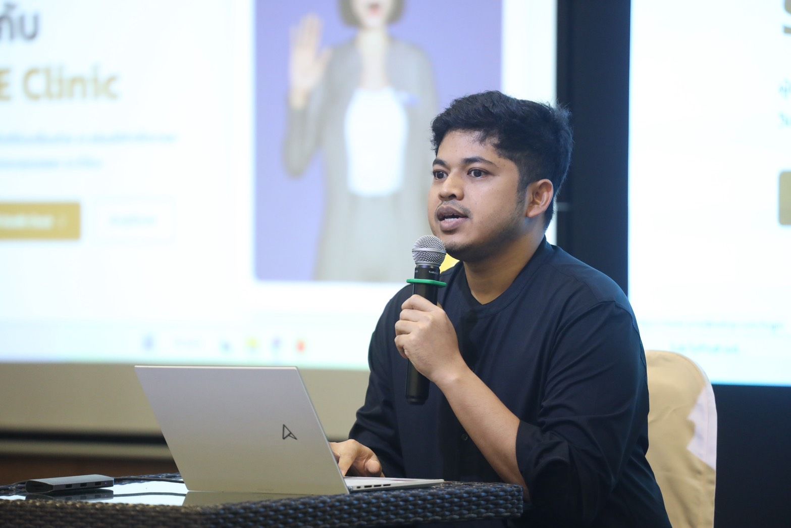 Young man speaks into microphone, laptop open, AI presentation screen in background.