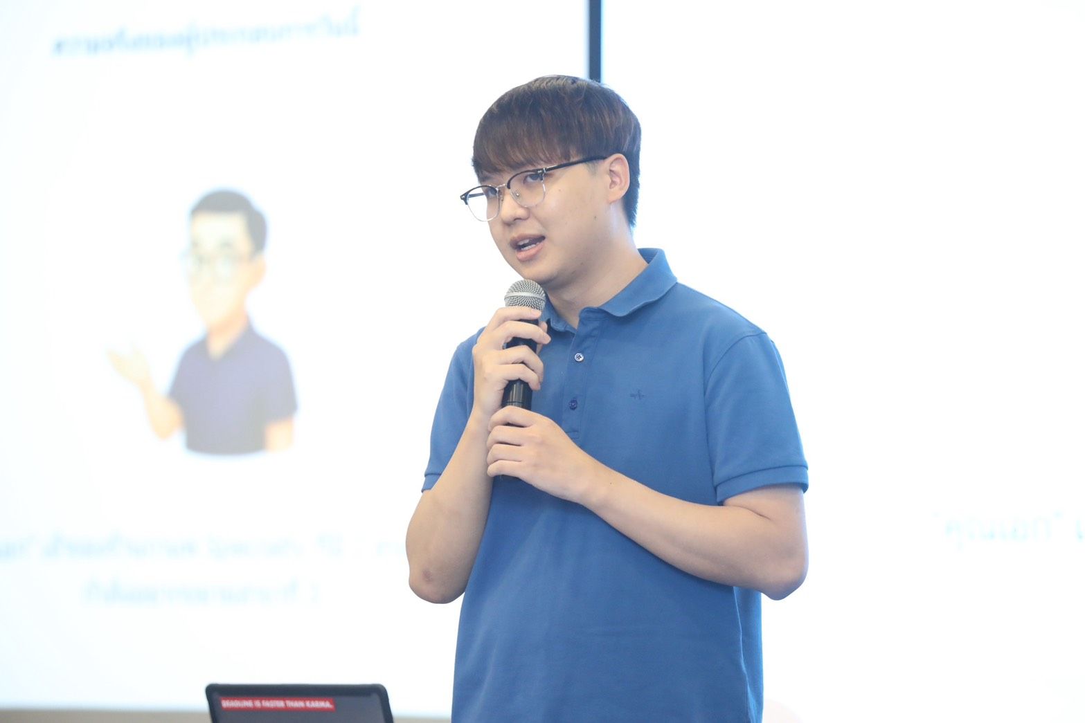 Man in blue polo shirt and glasses speaking into a microphone during a presentation.