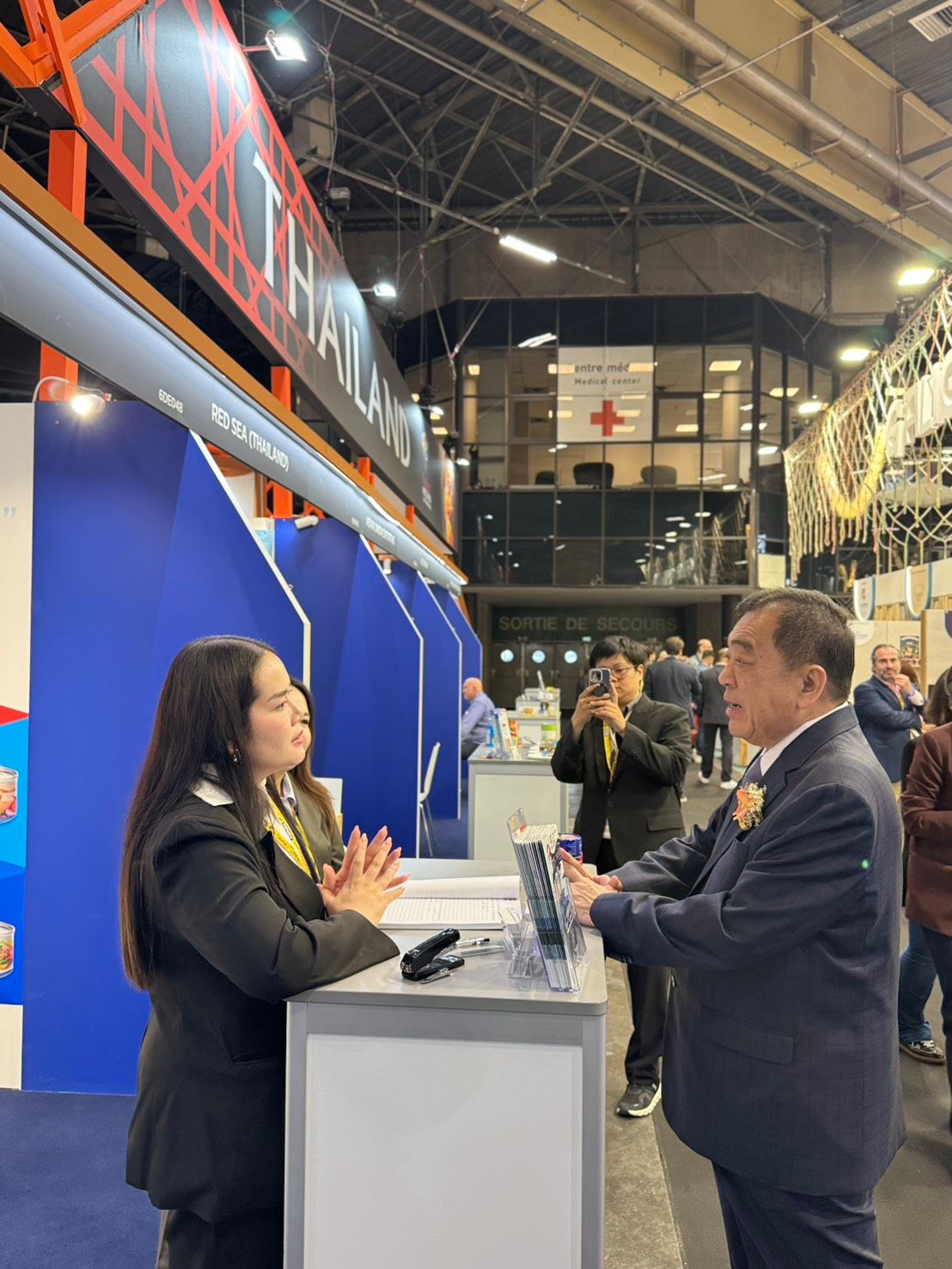 Two professionals converse at a blue exhibition booth in a large hall.