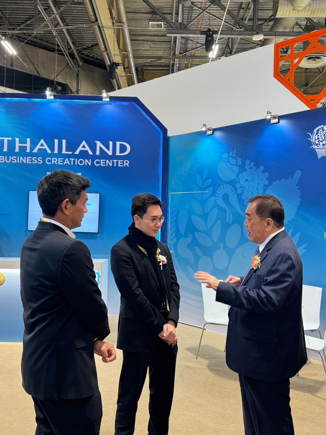 Three men in suits converse at a Thailand exhibition booth.