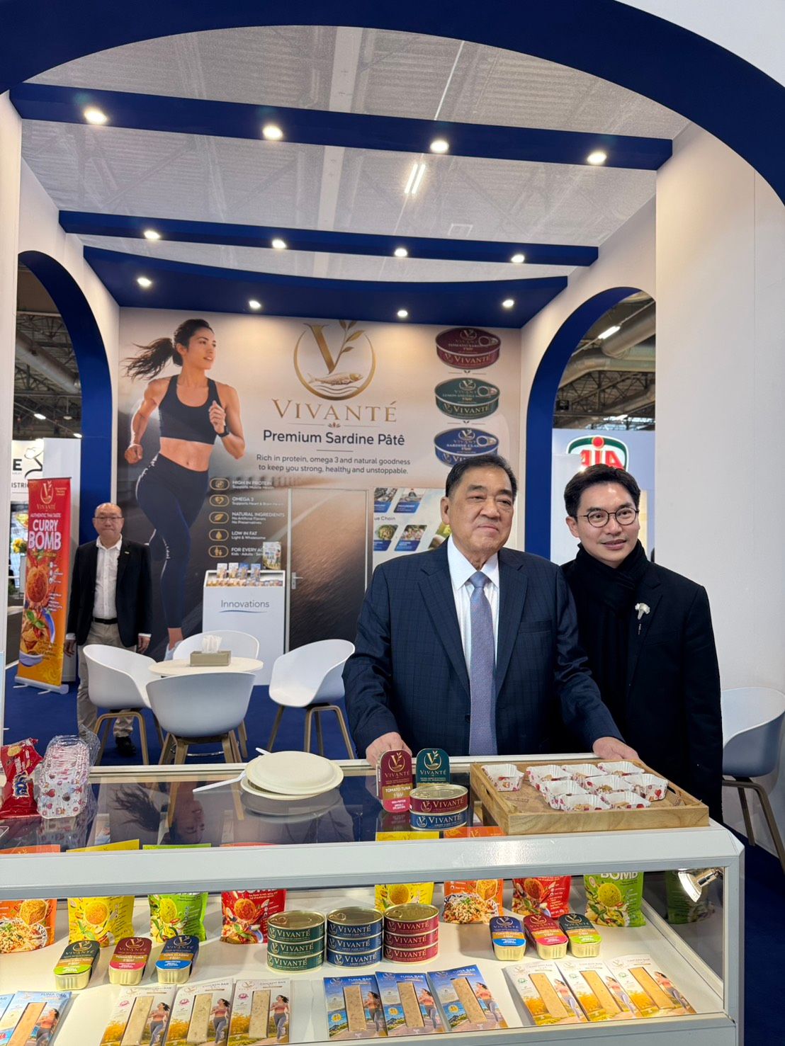 Two men stand at a food product booth, showcasing packaged items at a trade show.