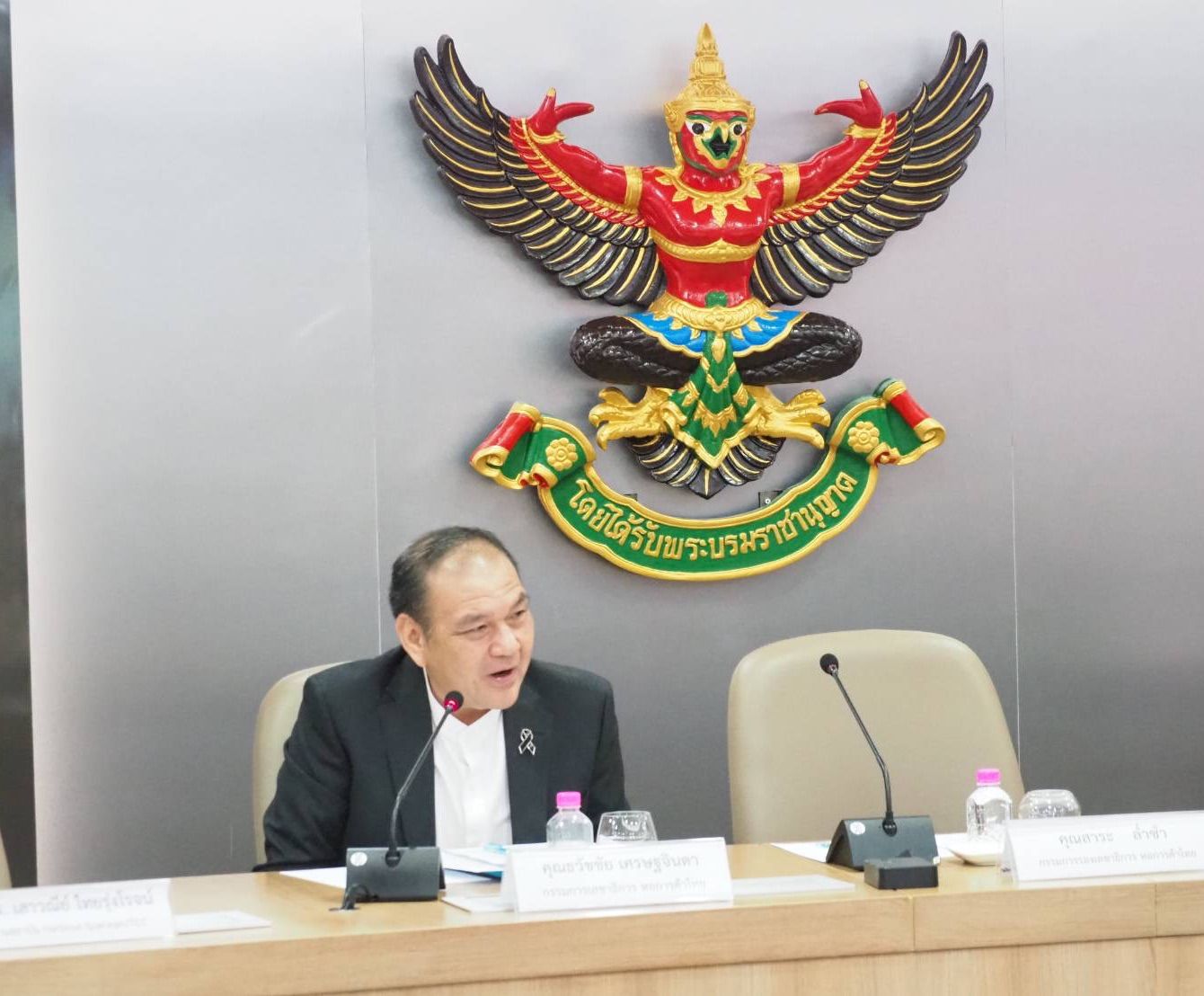 Man speaking at a table with a large golden Thai Garuda emblem behind him.
