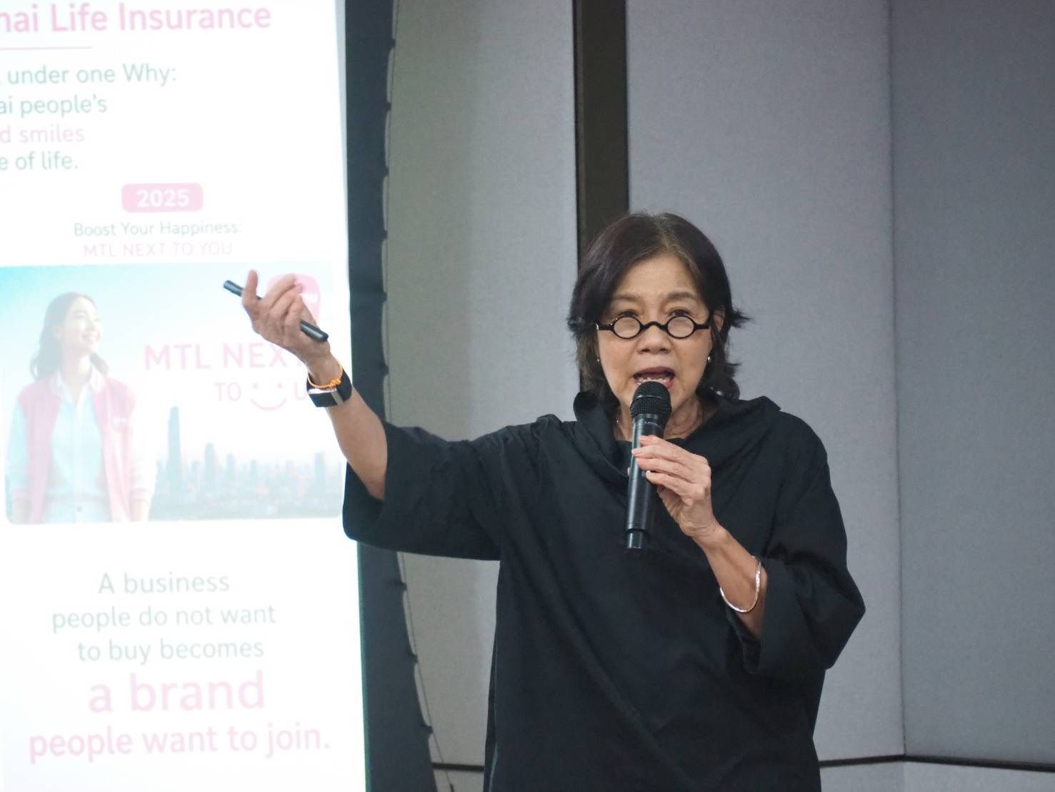 Woman in glasses speaks into a microphone, pointing at a presentation screen with text.