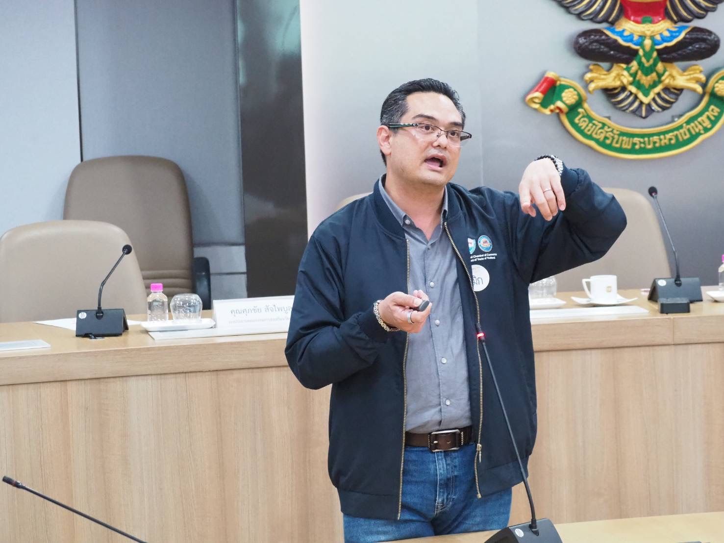 Man in dark jacket speaks and gestures during a conference room meeting.