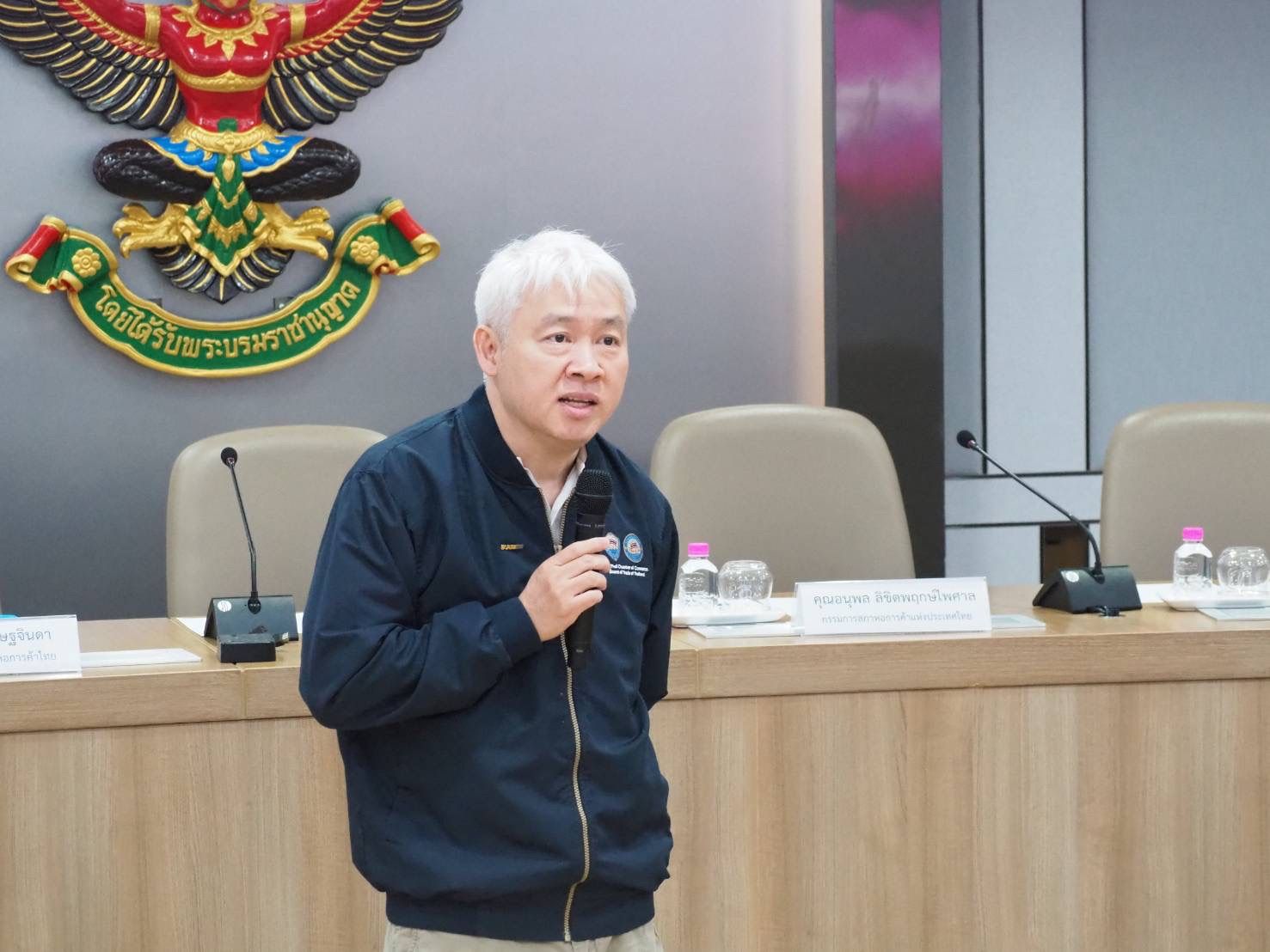 A white-haired man in a dark jacket speaks into a microphone at a formal presentation.