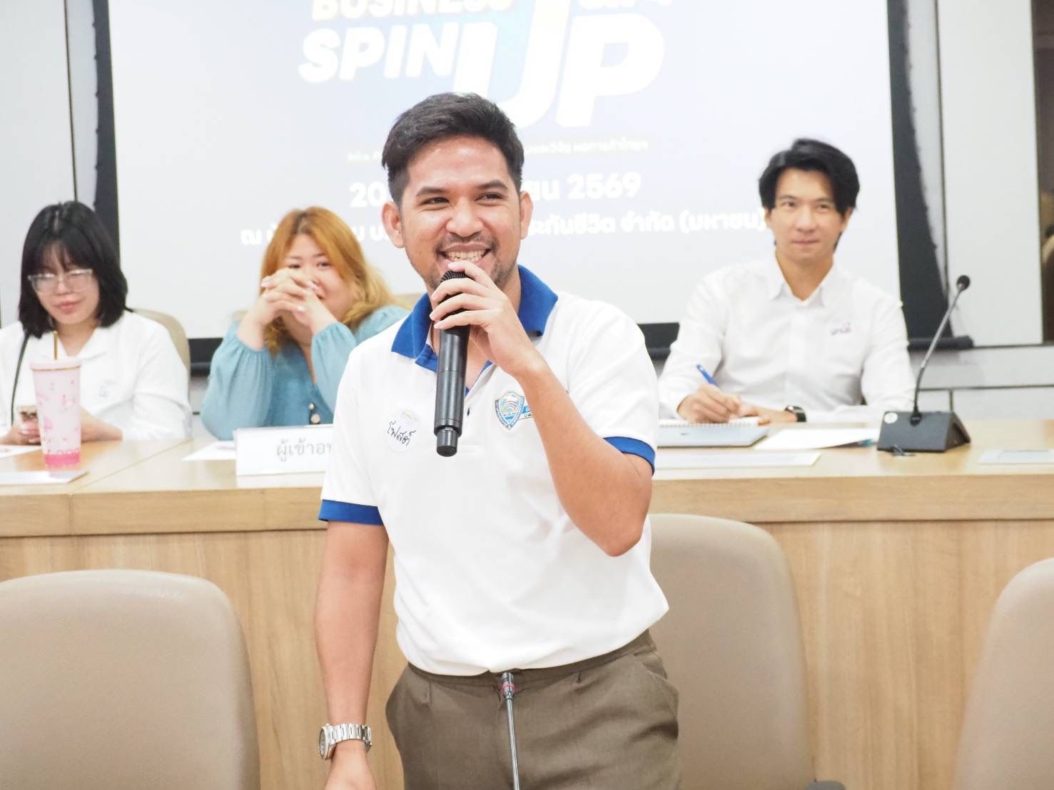 Smiling man in a white and blue polo shirt speaks into a microphone at a business event.
