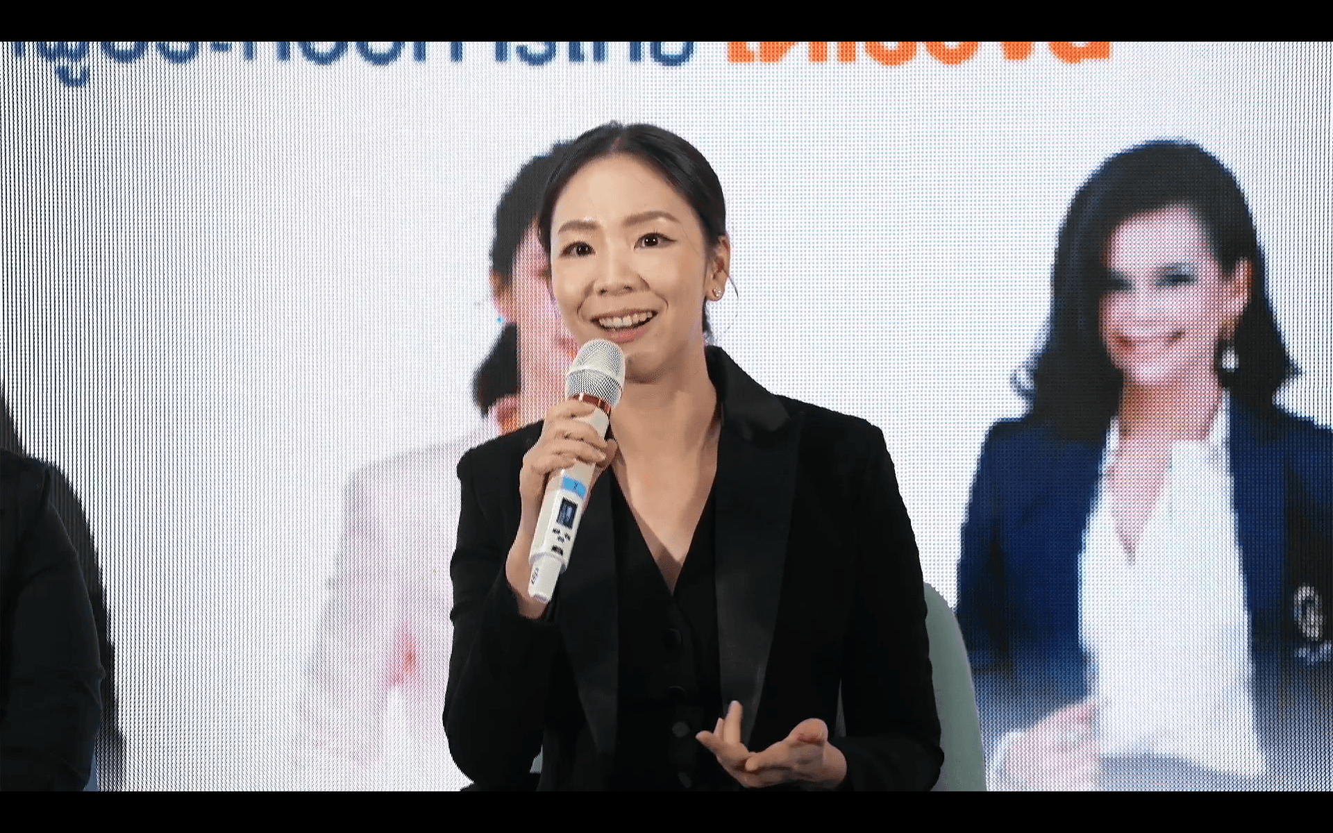 Smiling woman in a black blazer speaks confidently into a white microphone.