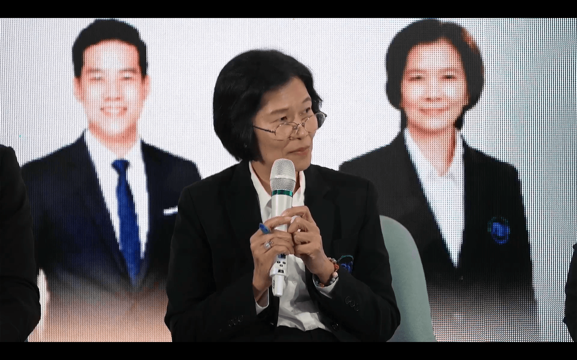 A woman in glasses speaks into a microphone at an event with blurred portraits behind her.