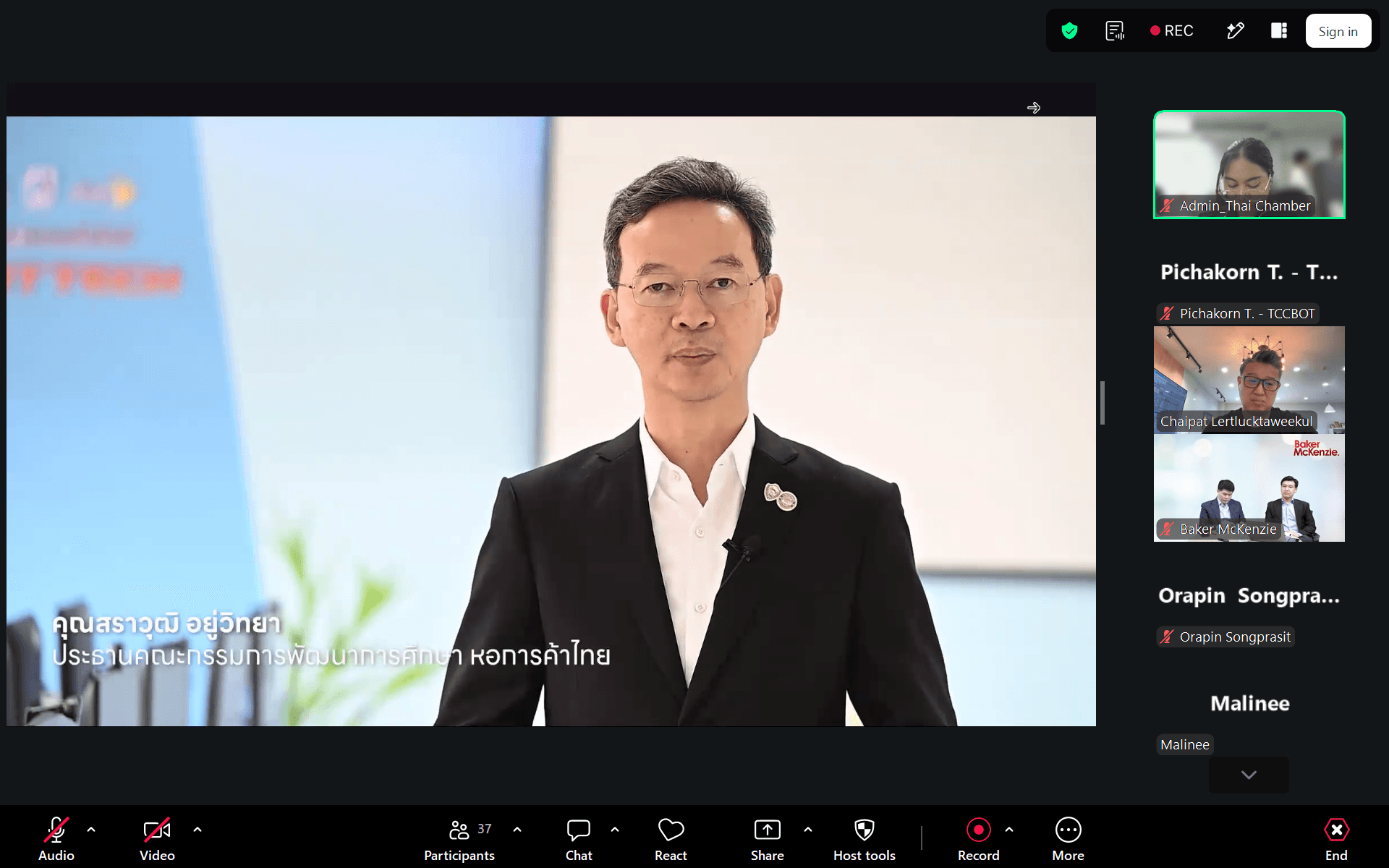 Man in a suit presenting during a virtual conference with multiple participants visible on screen.