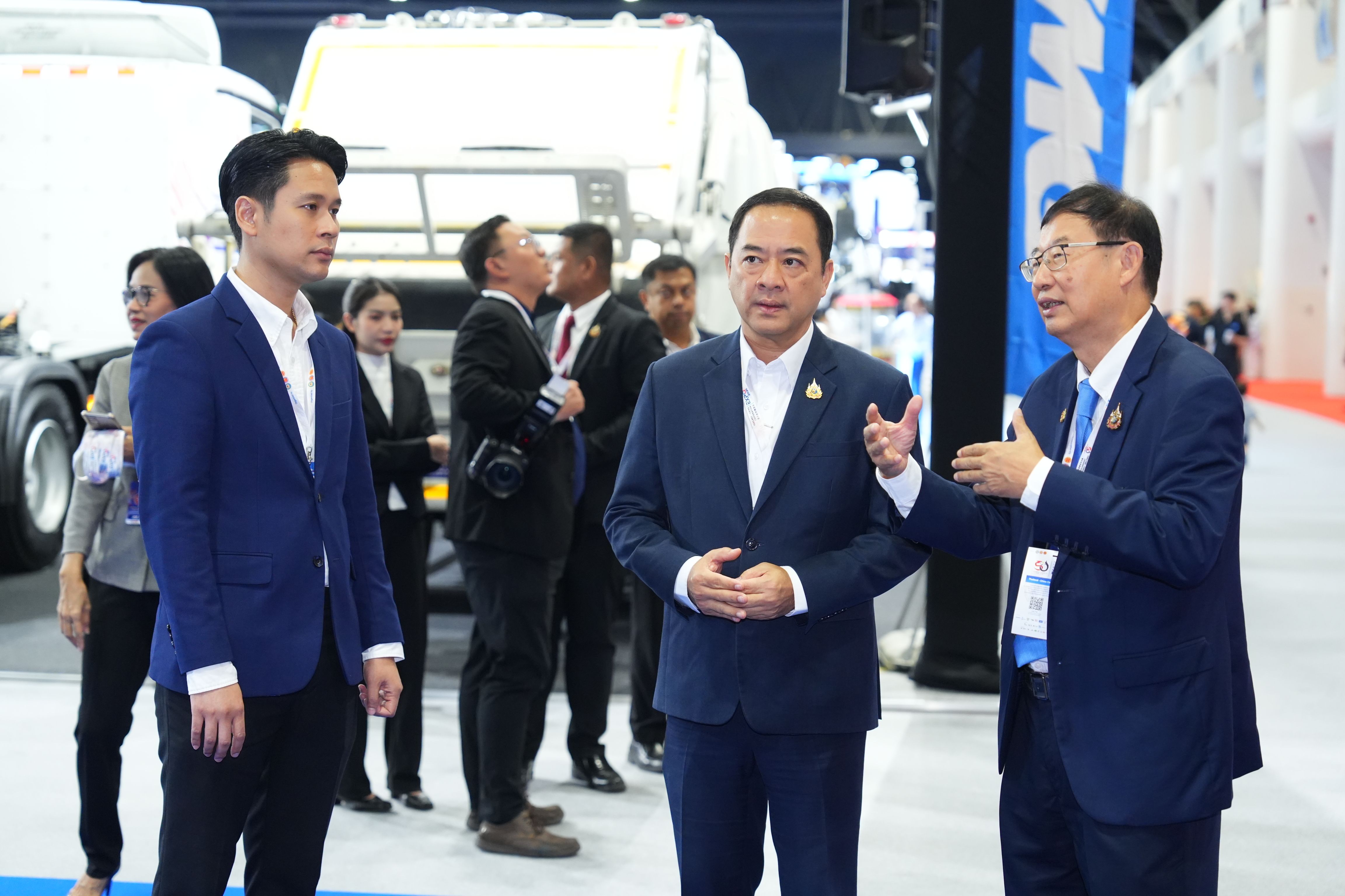 Three men in suits discuss at an exhibition, one gesturing towards another.