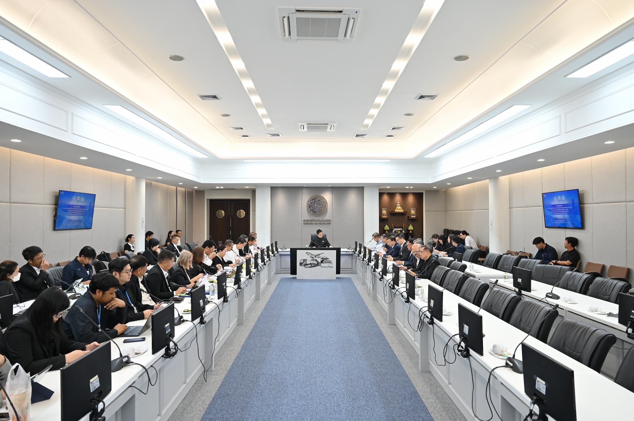 People attending a large formal meeting in a modern conference room with monitors.