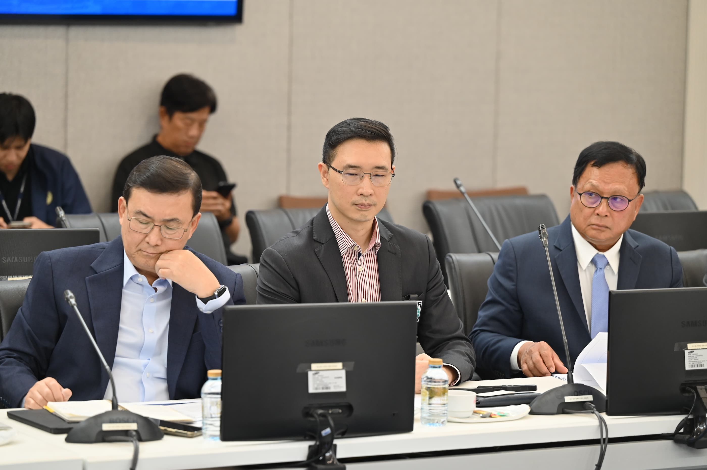 Three men in business suits attend a meeting at a conference table with laptops.