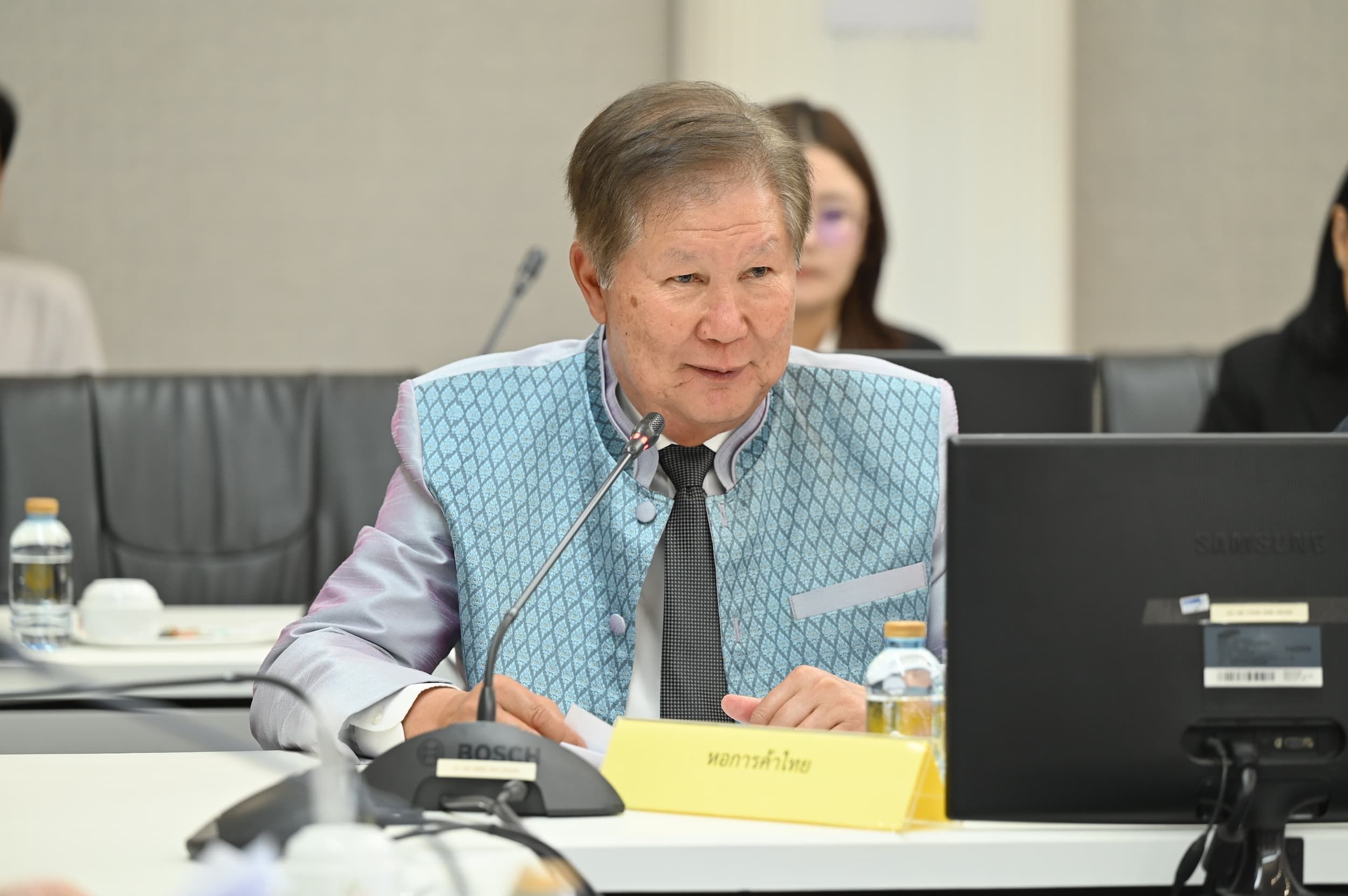 Elderly man in a patterned blue shirt sits at a conference table, speaking.