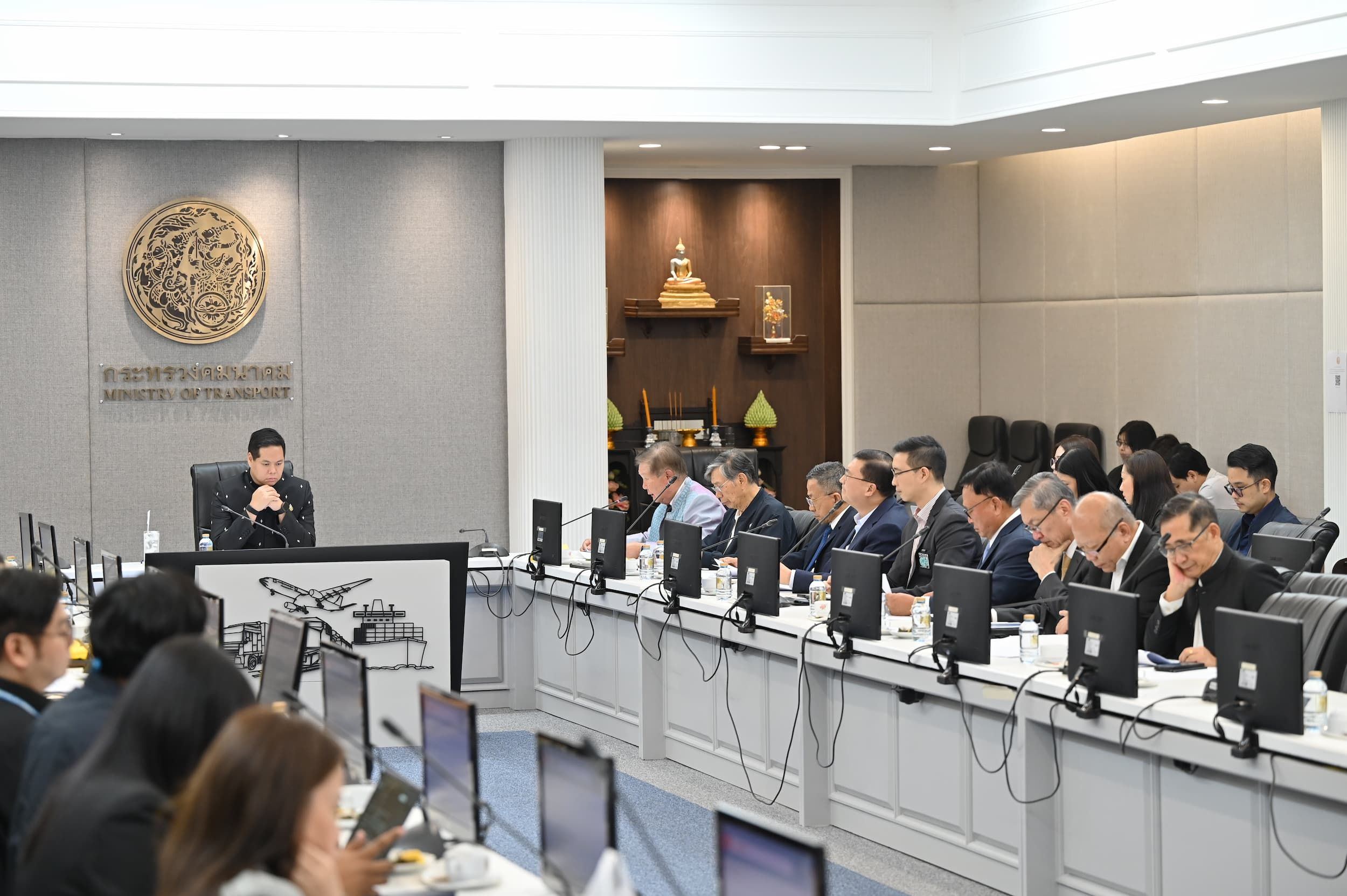 A person addresses a meeting of officials seated at a long conference table with monitors.