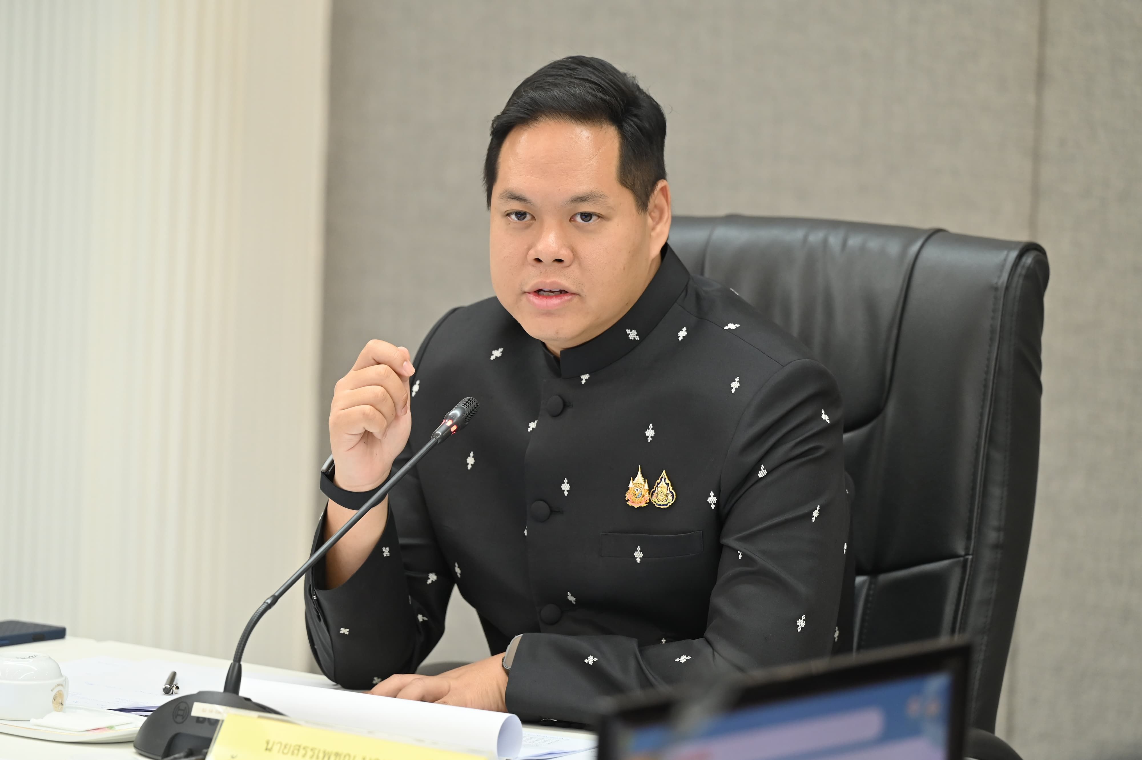 A man in a dark, embroidered jacket speaks at a formal meeting with a microphone.