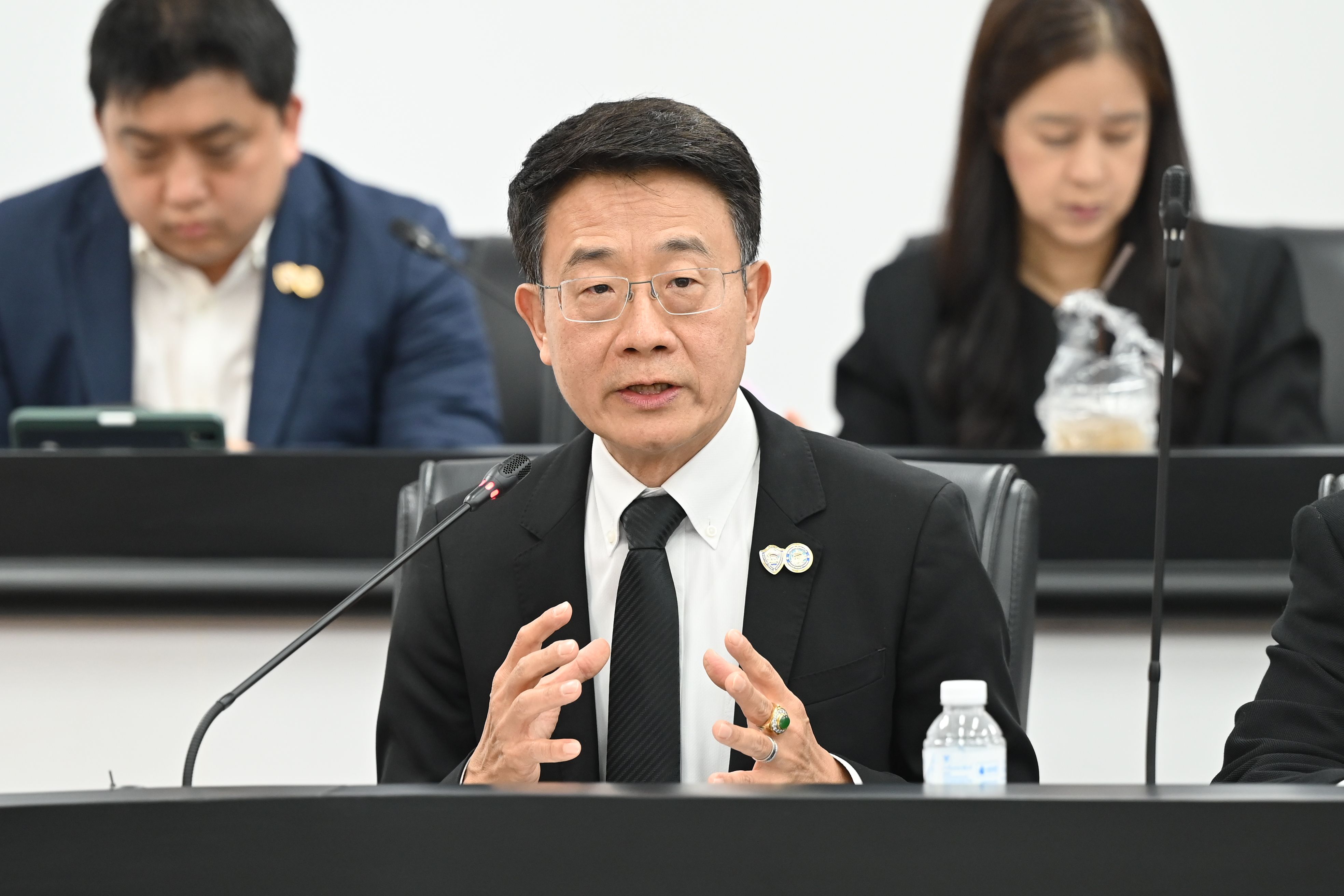 A man in a dark suit and glasses speaks at a table, gesturing with both hands.