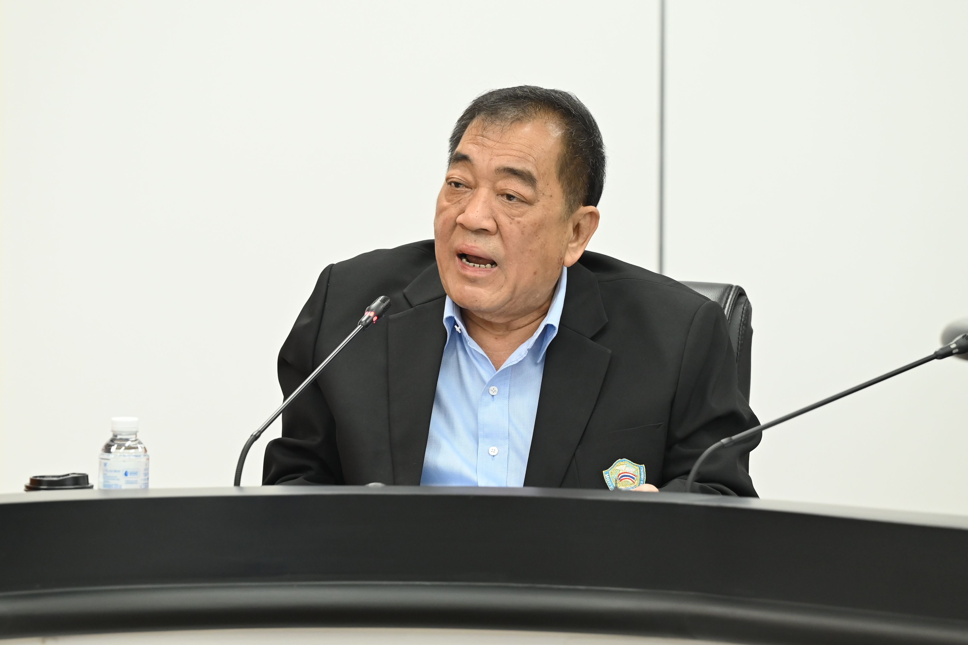Man in a dark suit jacket and blue shirt speaking into microphones at a table.