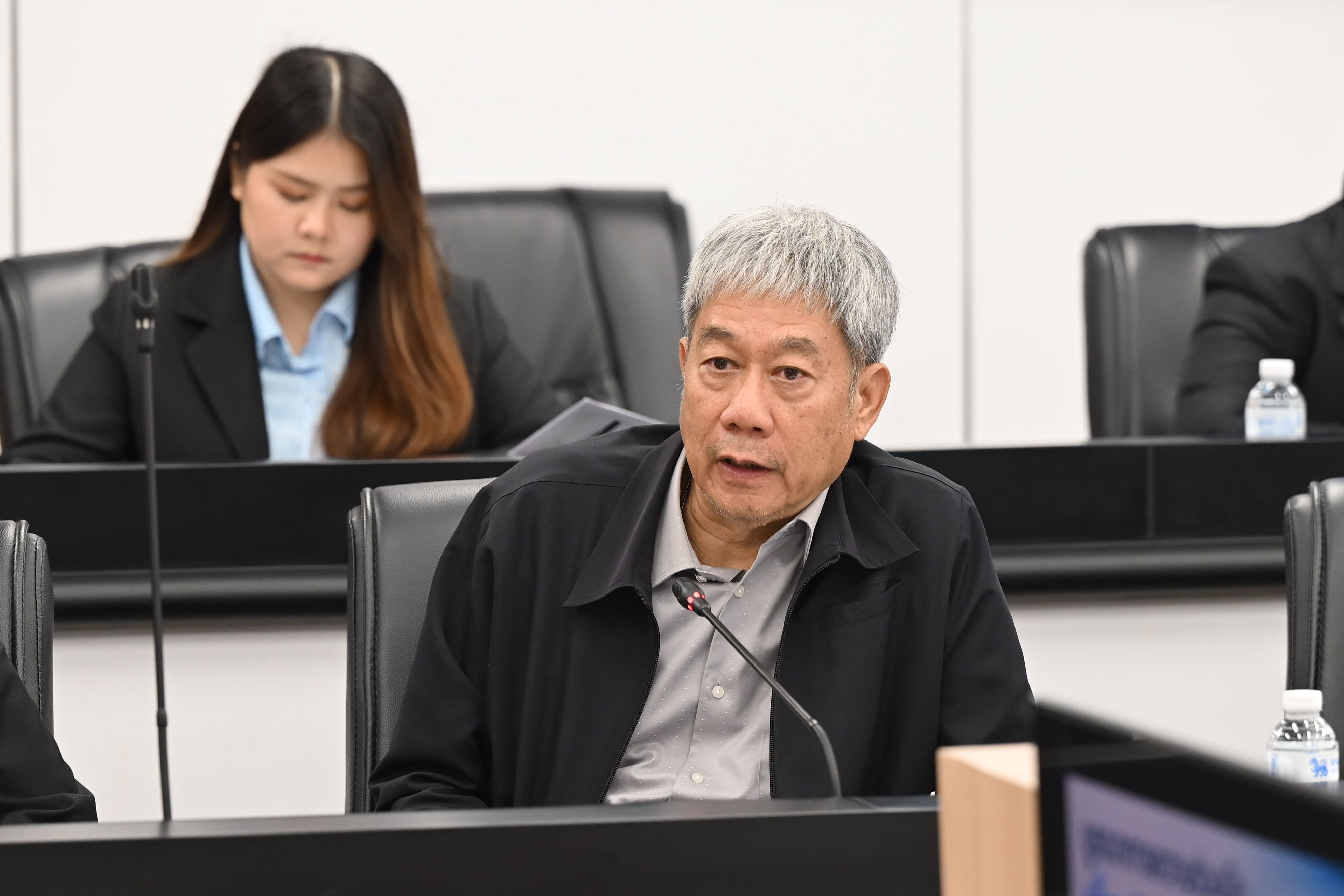 Elderly Asian man with grey hair speaking intently into a microphone at a formal meeting.