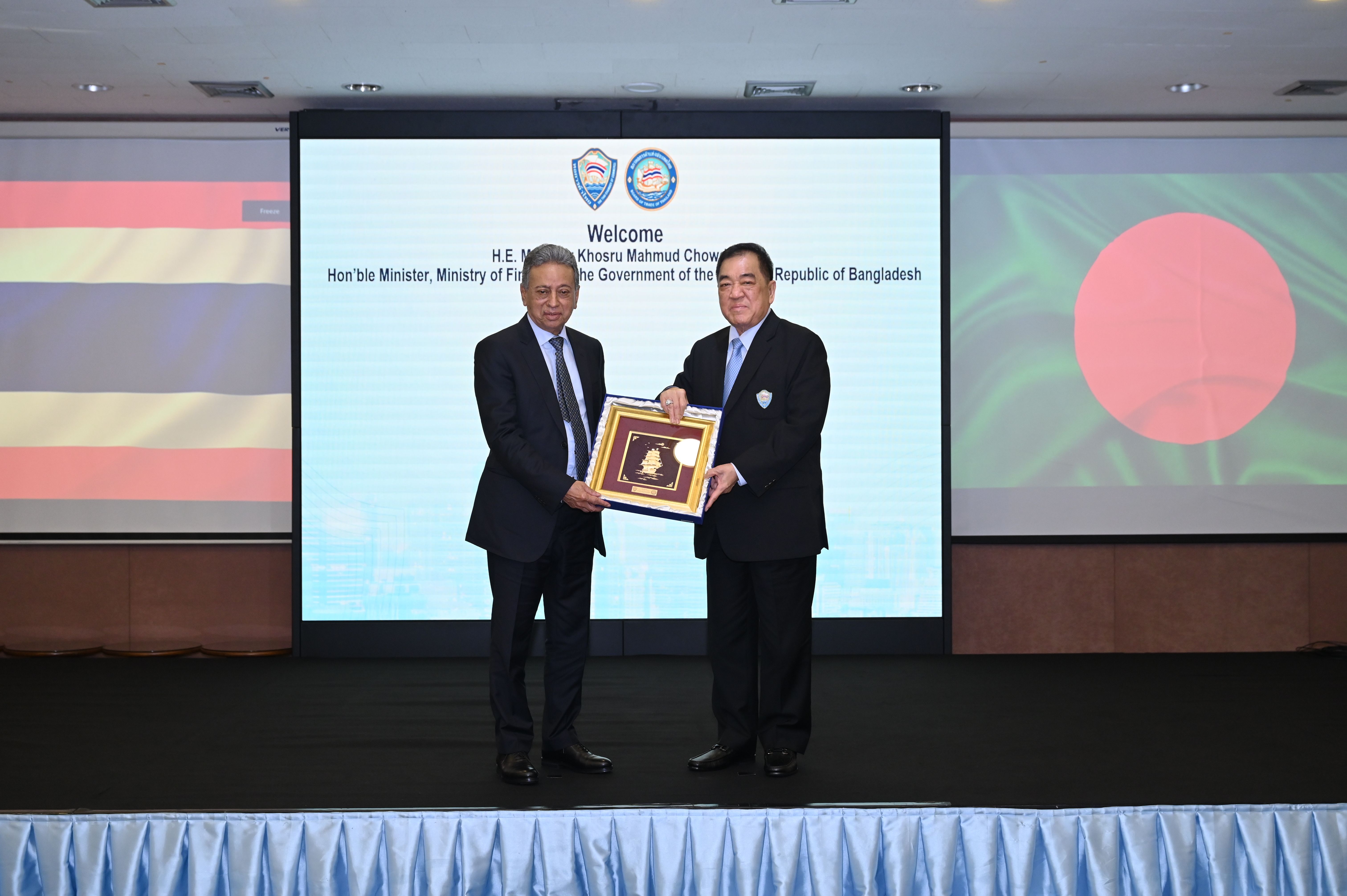 Two men in suits exchange a framed gift on stage with a 'Welcome' screen behind them.