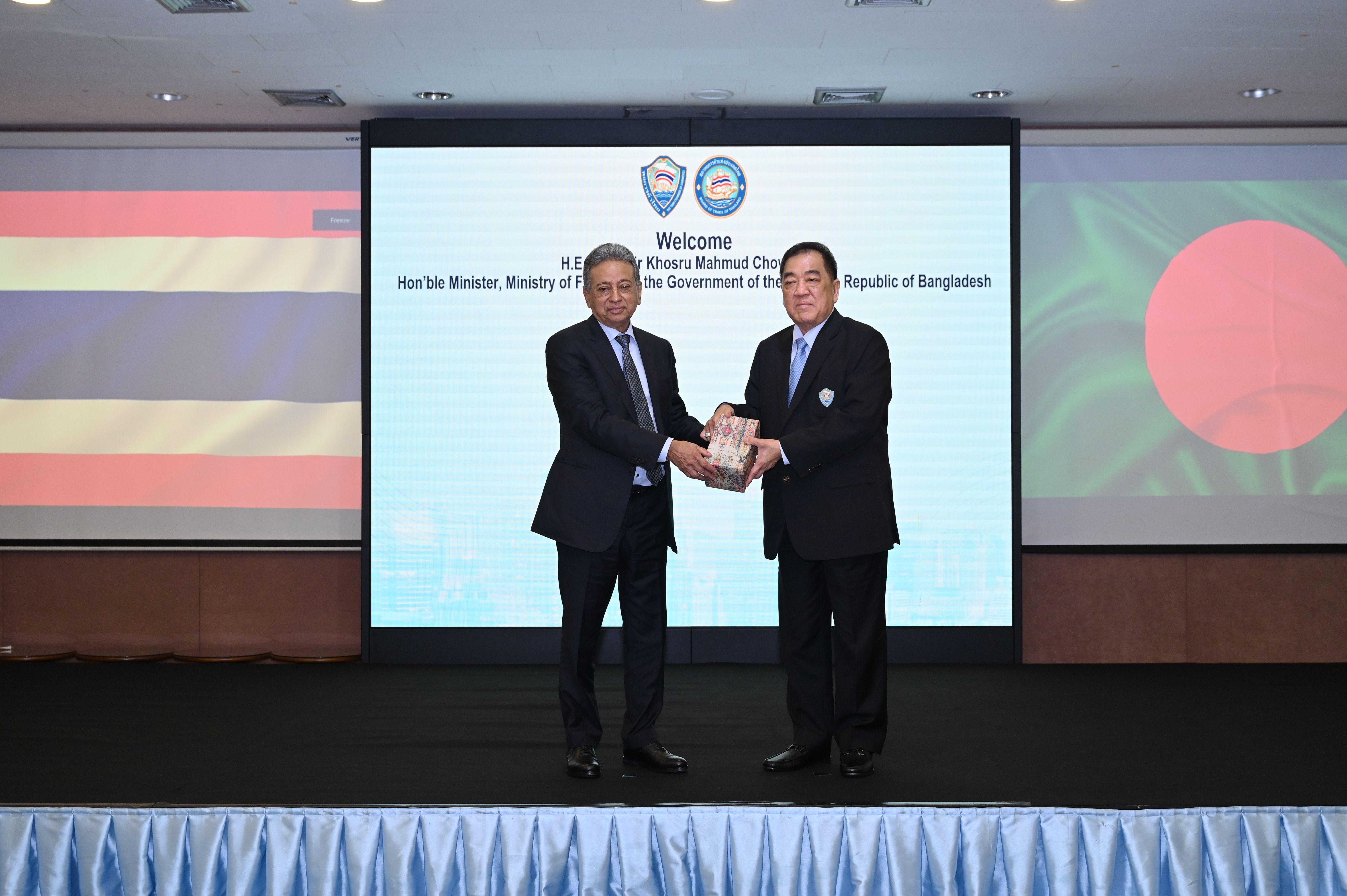 Two formally dressed men on stage exchanging a small item, with flags and a welcome screen behind them.