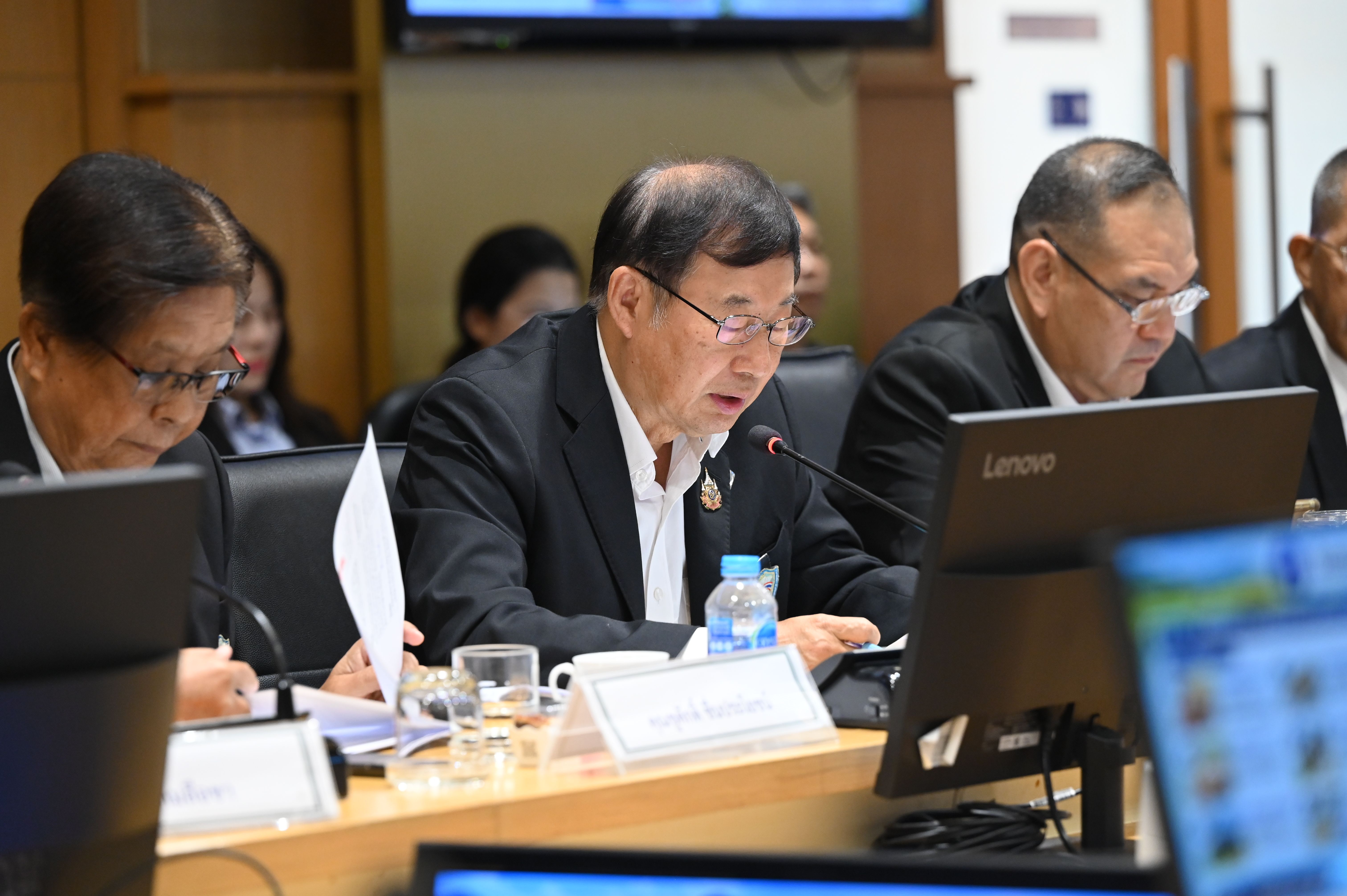 Three men attend a conference, one actively speaking into a microphone at the table.