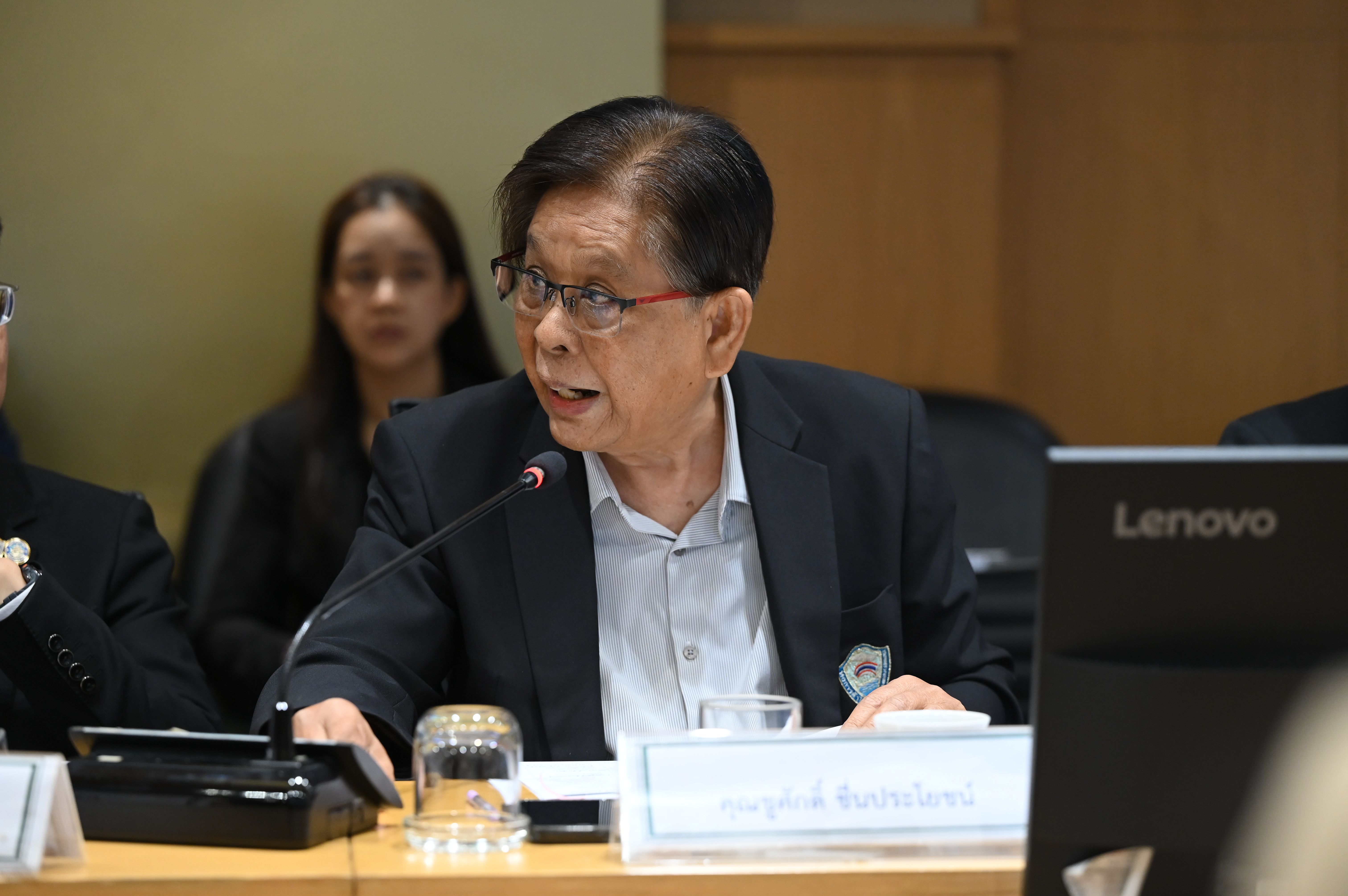 An older man in glasses and a suit speaks at a conference table with a microphone.