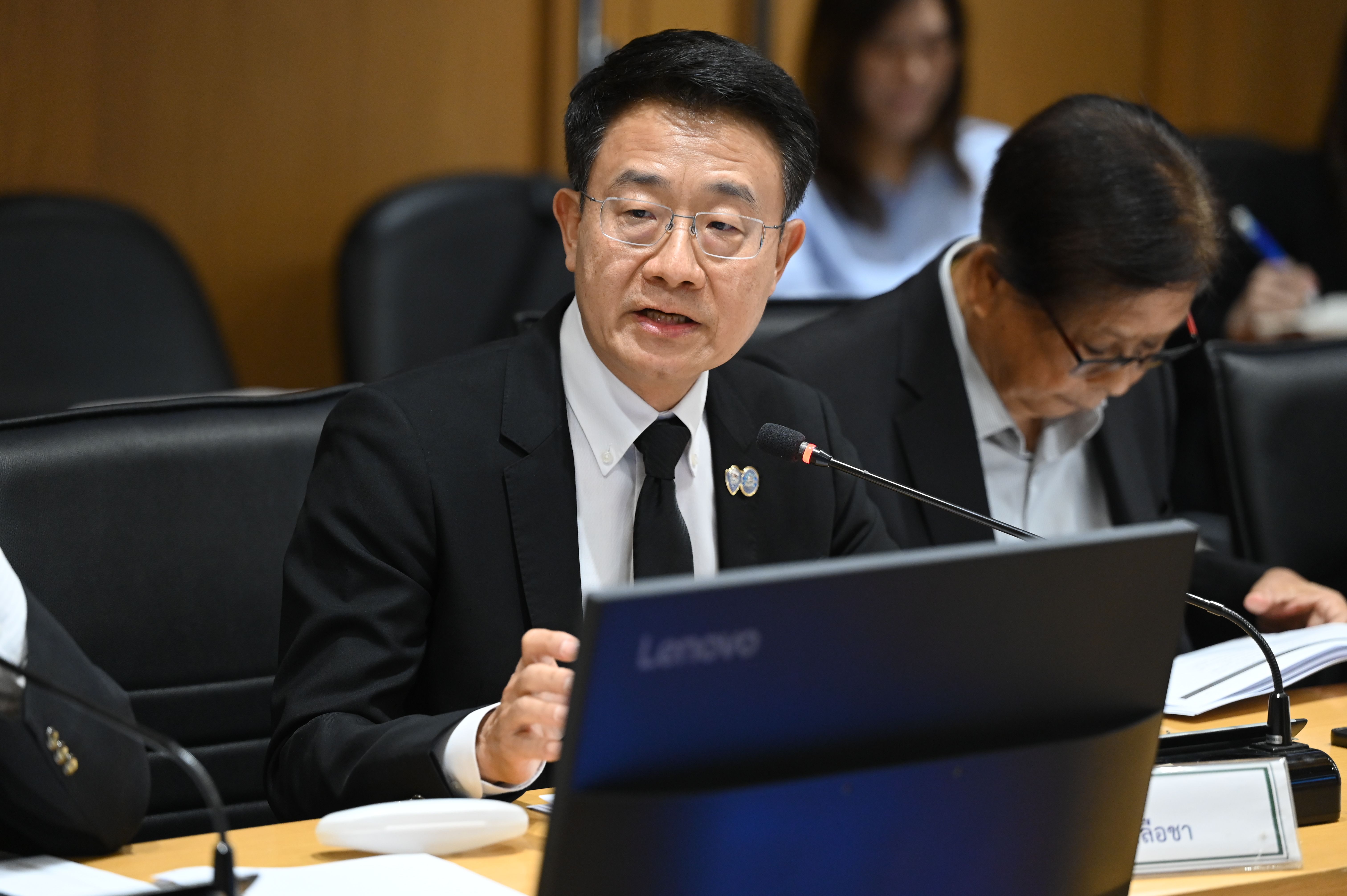 Man in suit speaks at a meeting, gesturing with hands.