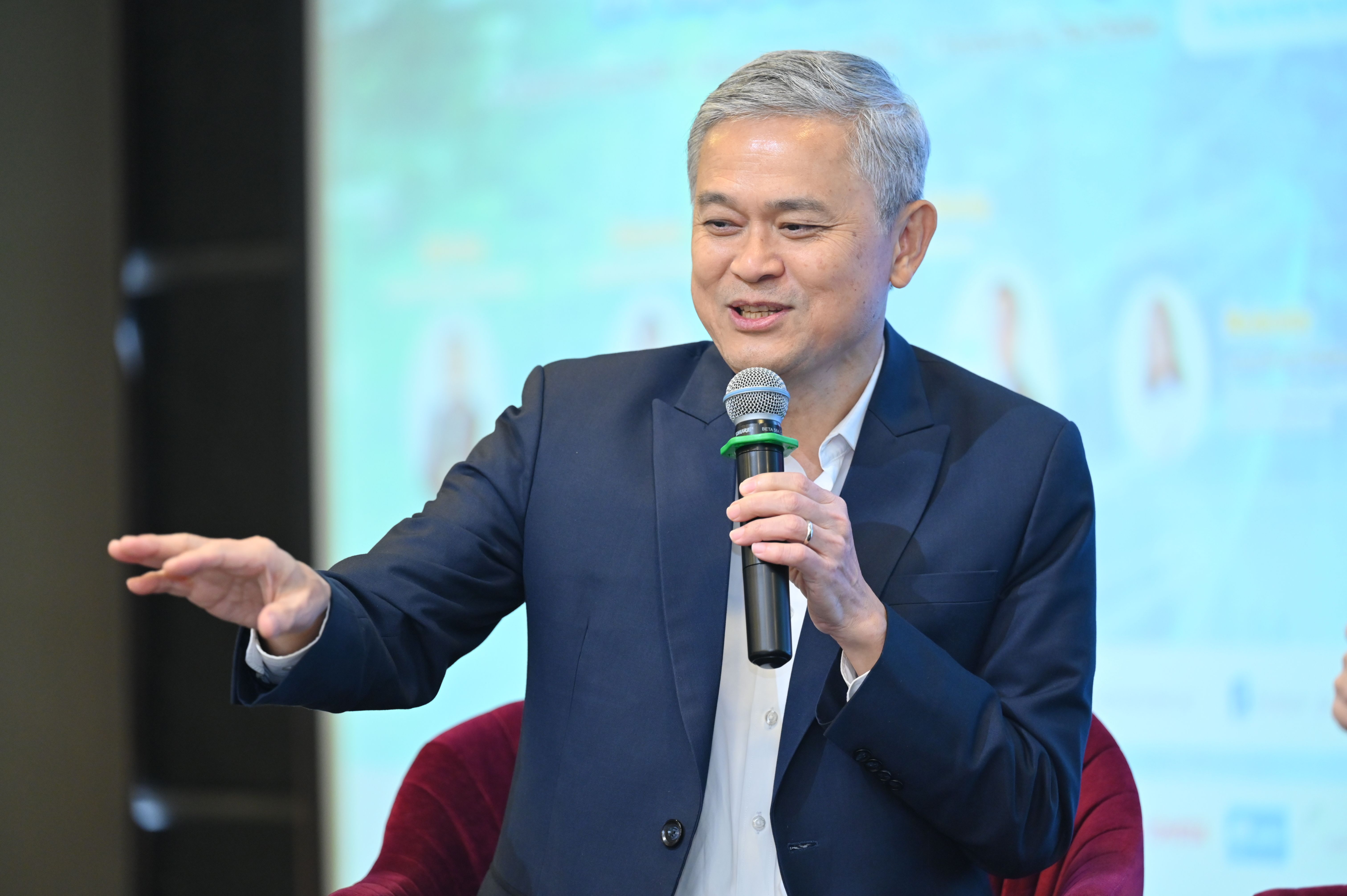 Grey-haired man in a dark suit speaks into a microphone, gesturing with his hand.