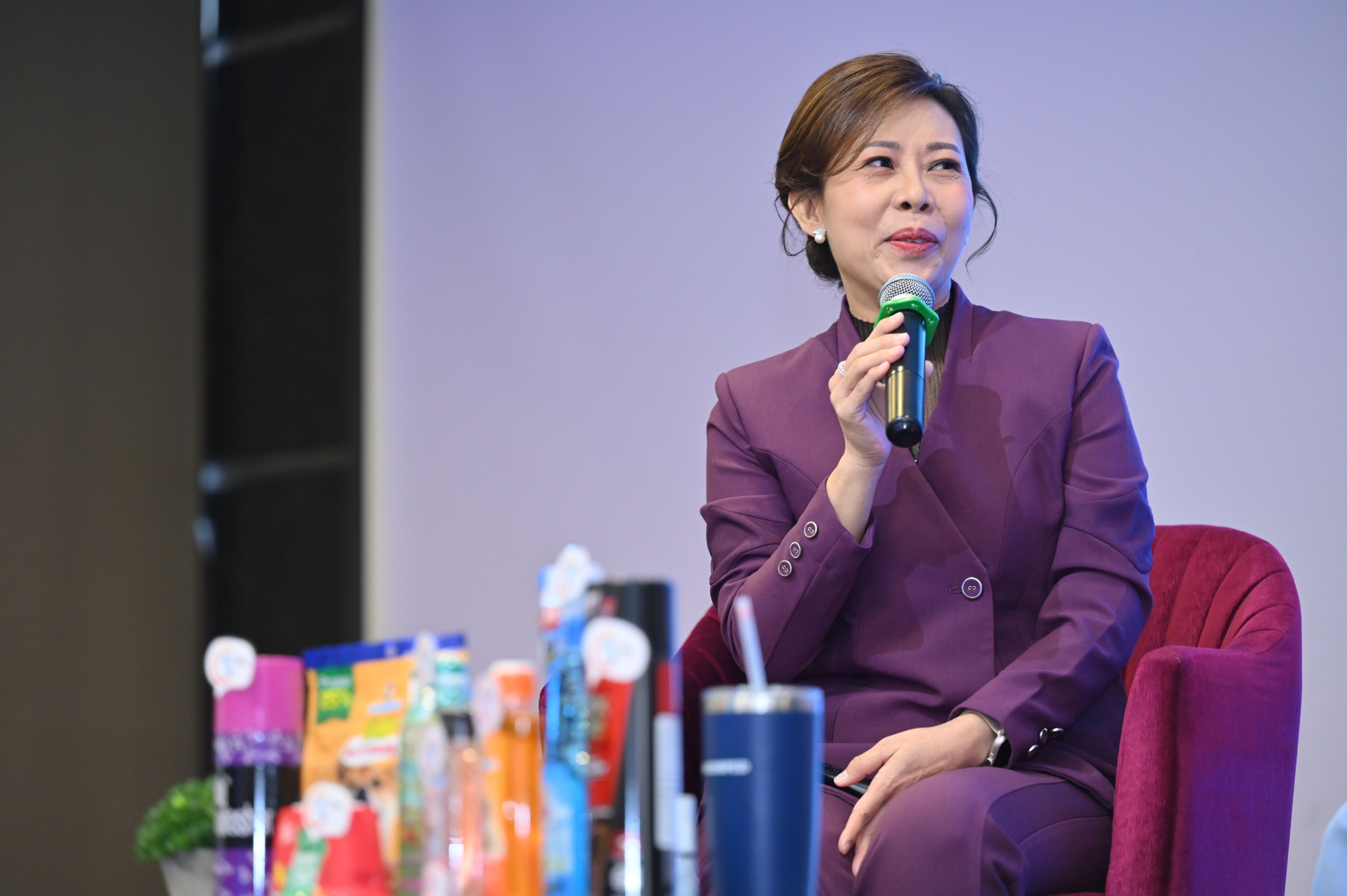 A professional woman in a purple suit speaks into a microphone at an event.