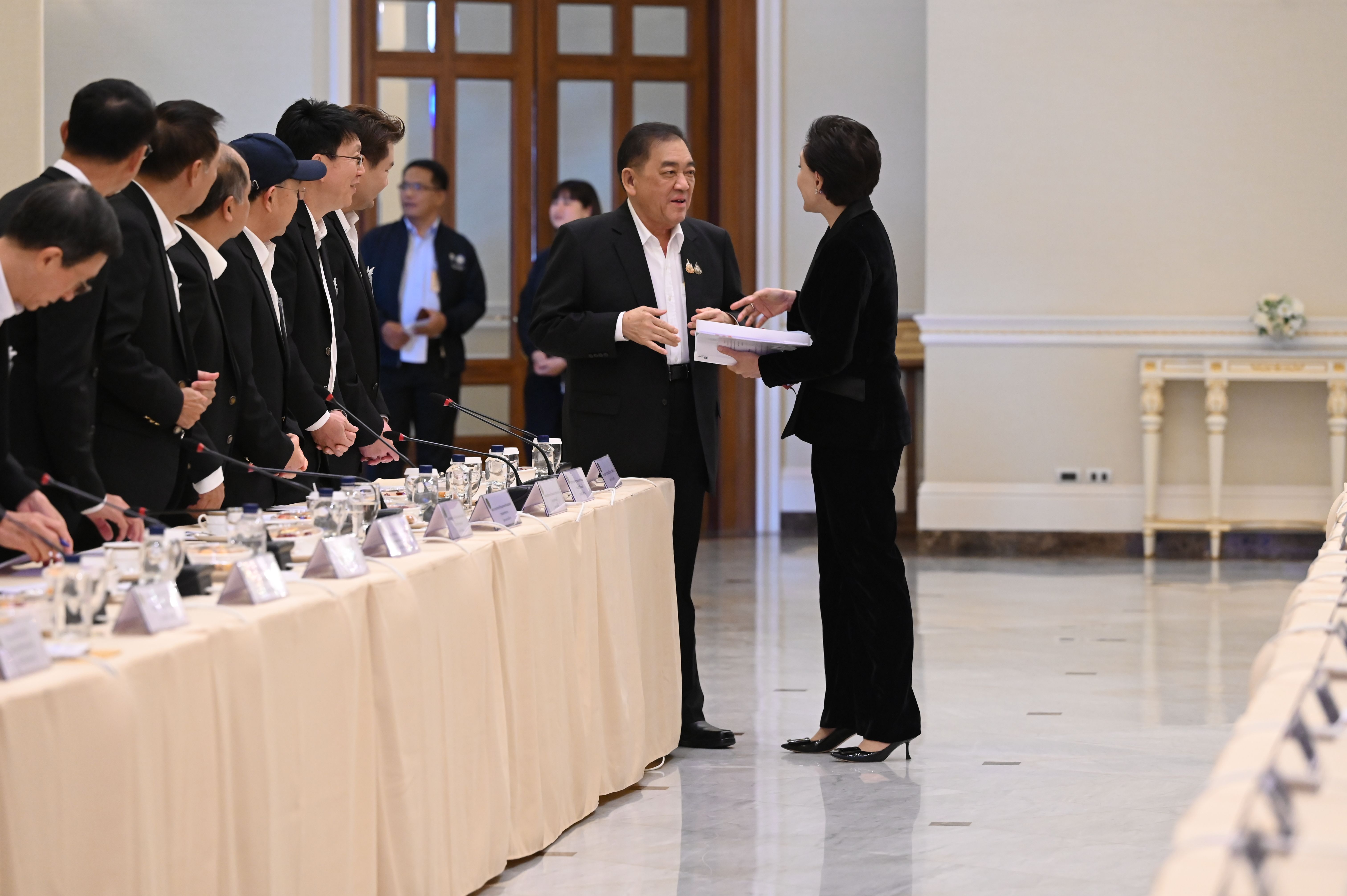 A man hands documents to a woman at a formal meeting table with other attendees.