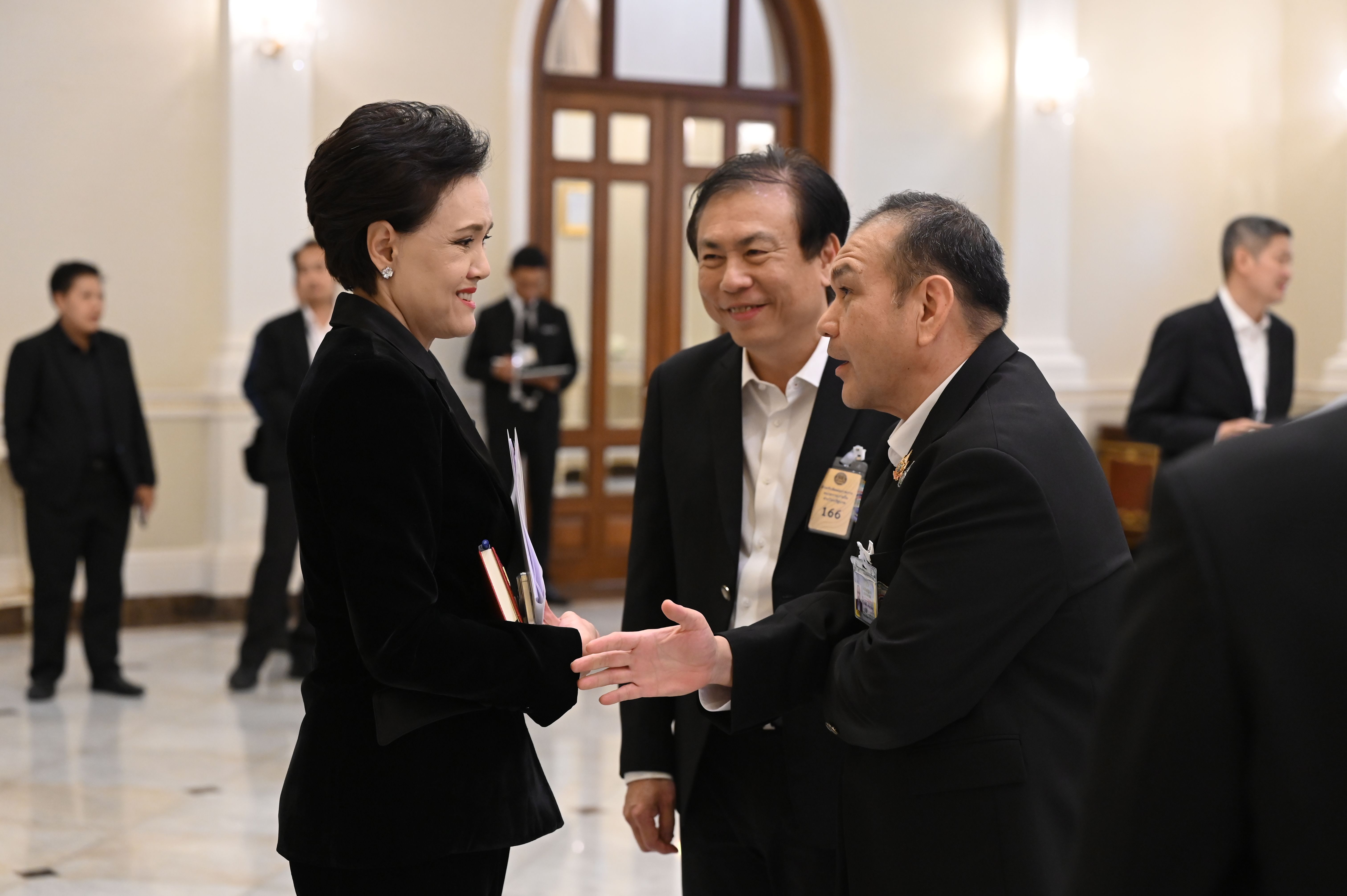 Three formally dressed people, a woman and two men, interact in a grand hall.
