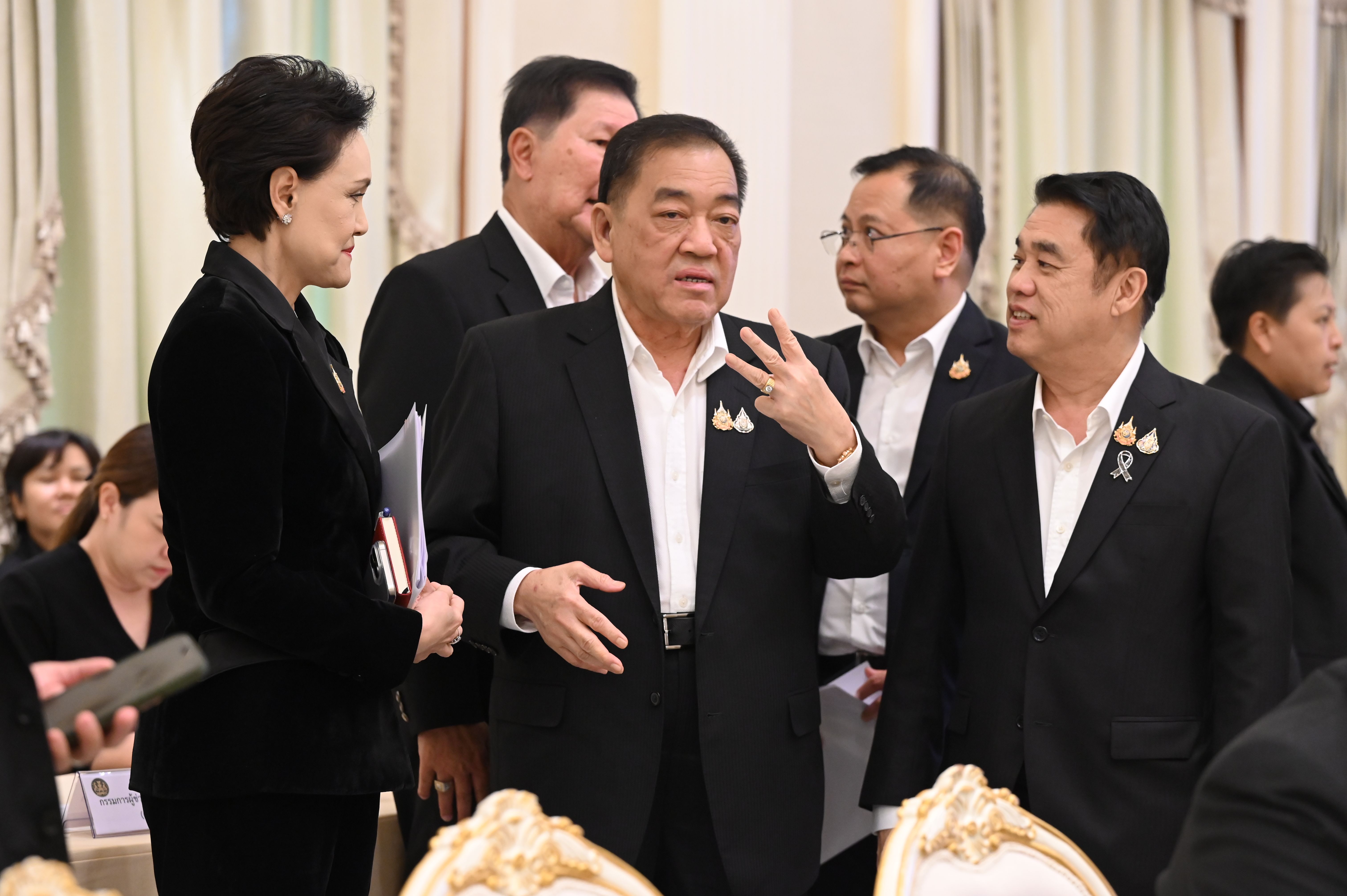 A man in a dark suit gestures while speaking to a group of colleagues.