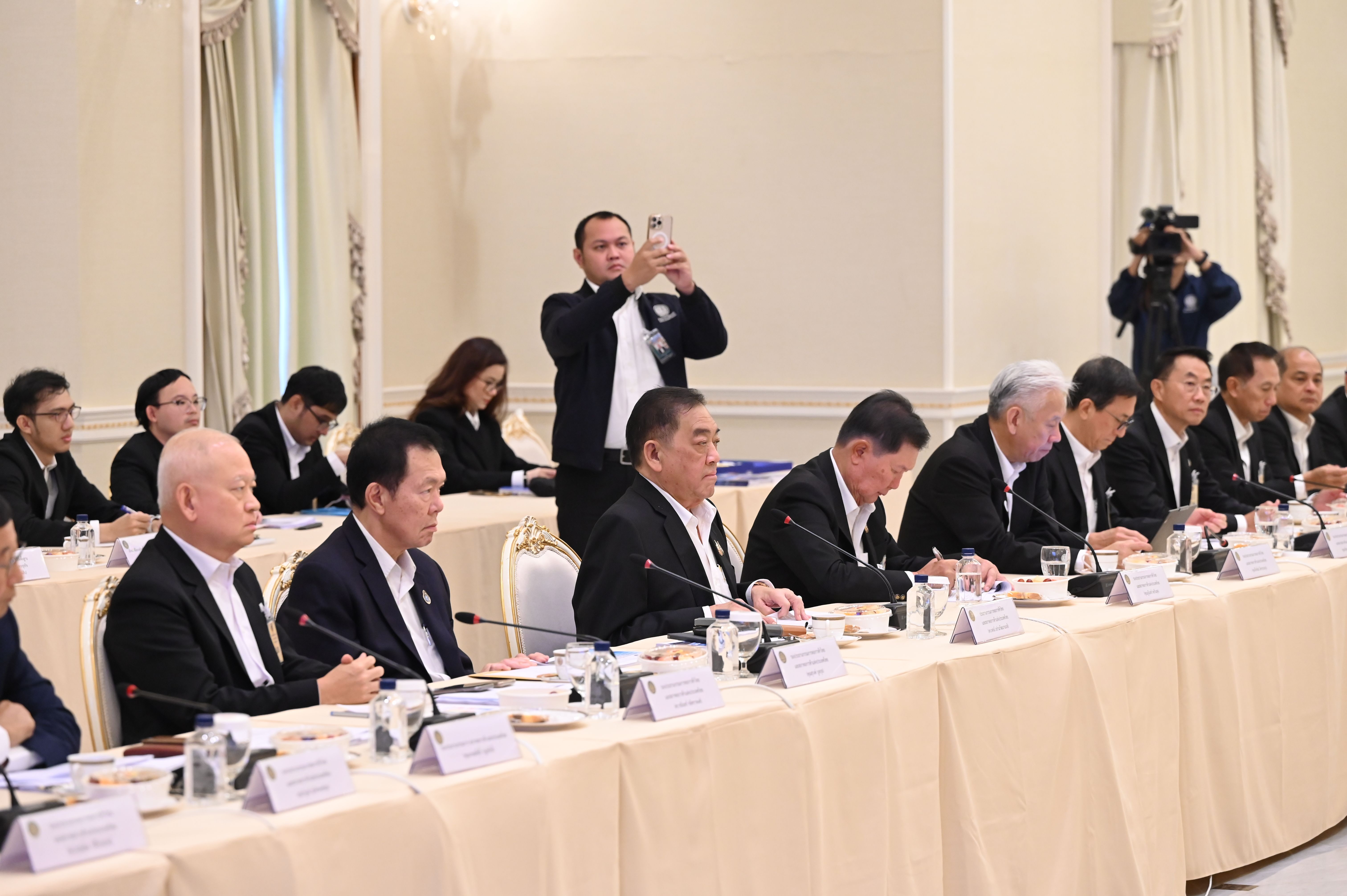 Men in dark suits attend a formal meeting at a long table indoors.